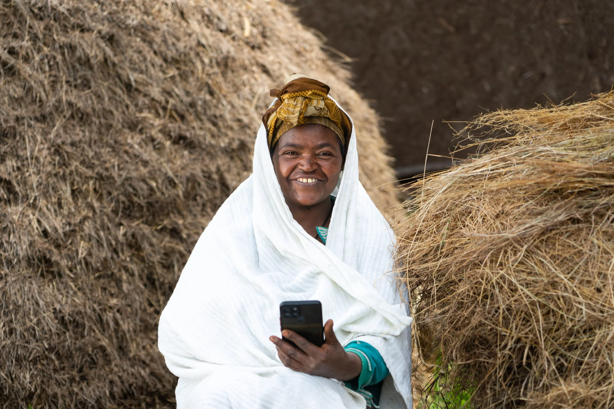 Smiling woman sitting between haystacks, holding a smartphone, wearing a white cloth and a headwrap.