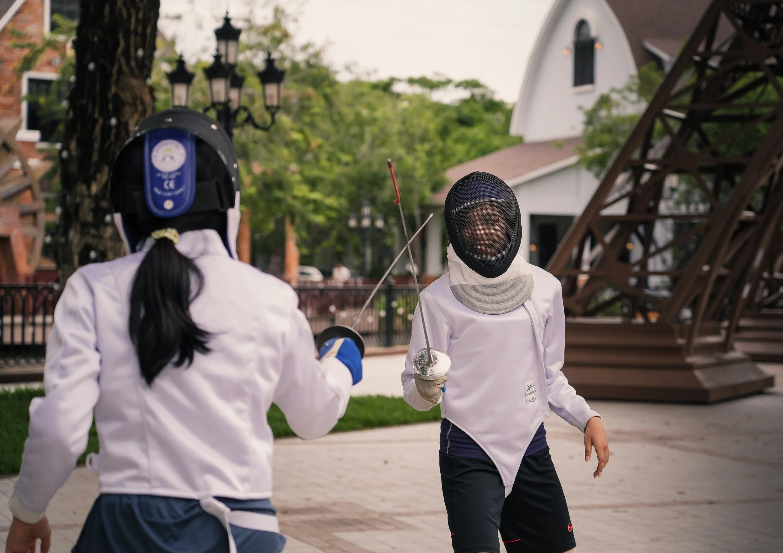 Two young women wearing fencing masks and fencing gear engaged in a fencing duel outdoors, with one woman holding a fencing foil.