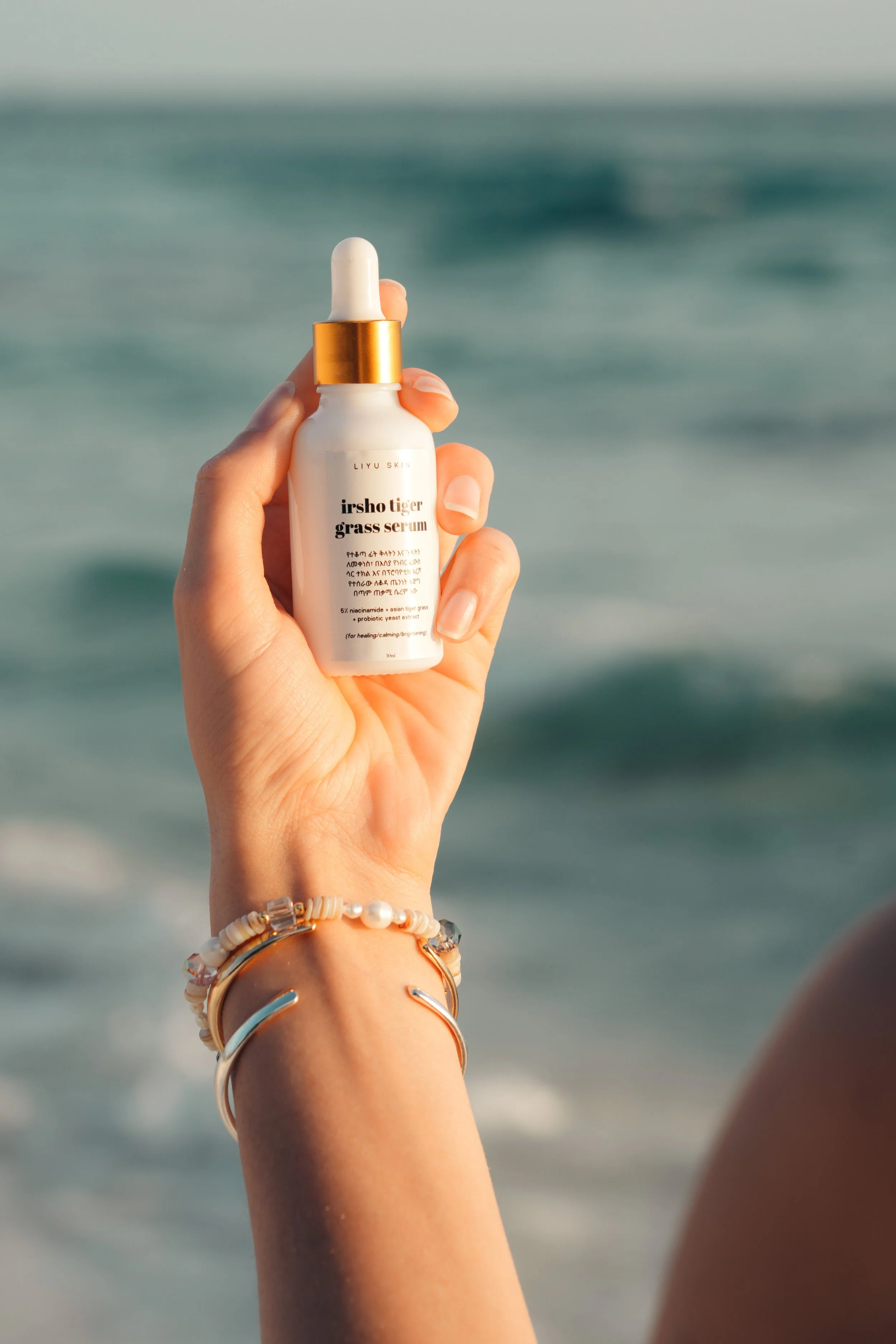 A hand holding a white bottle of skincare serum with a gold cap and dropper, against a blurred ocean background.