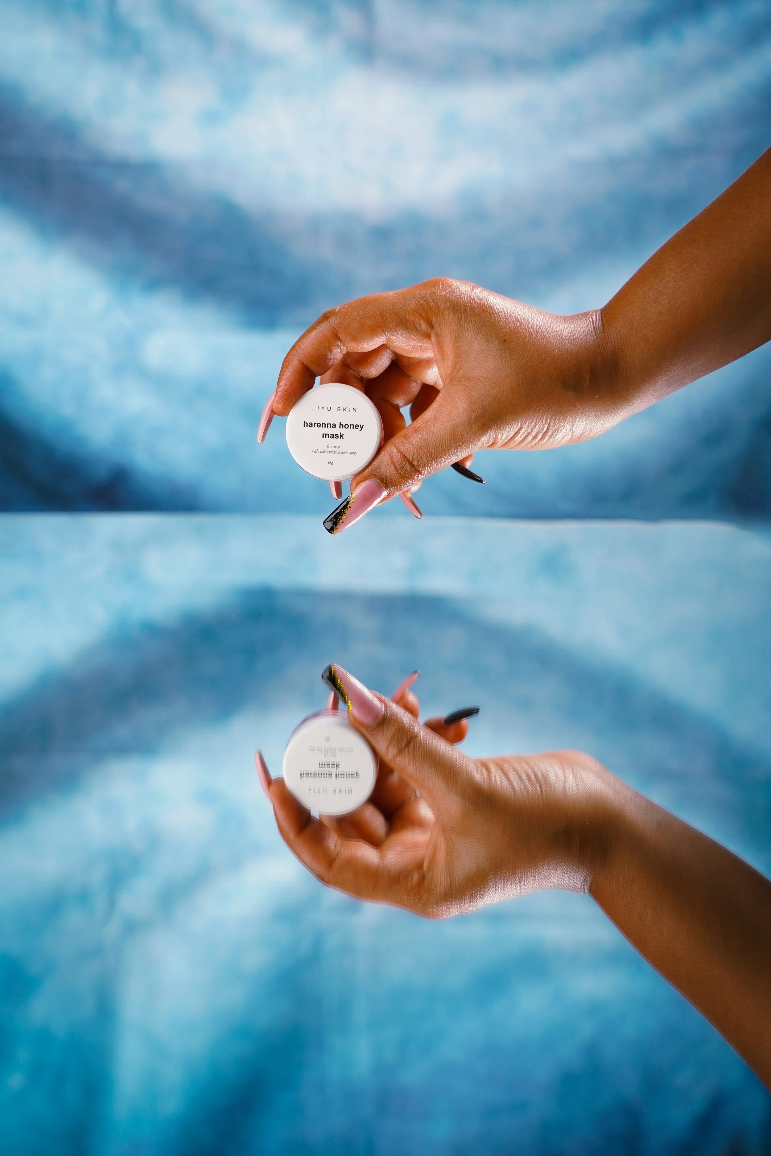 A person's hand holding a small white jar labeled 'LIYU SKIN hareuna honey mask' with a blue abstract background. reflected in a mirror.