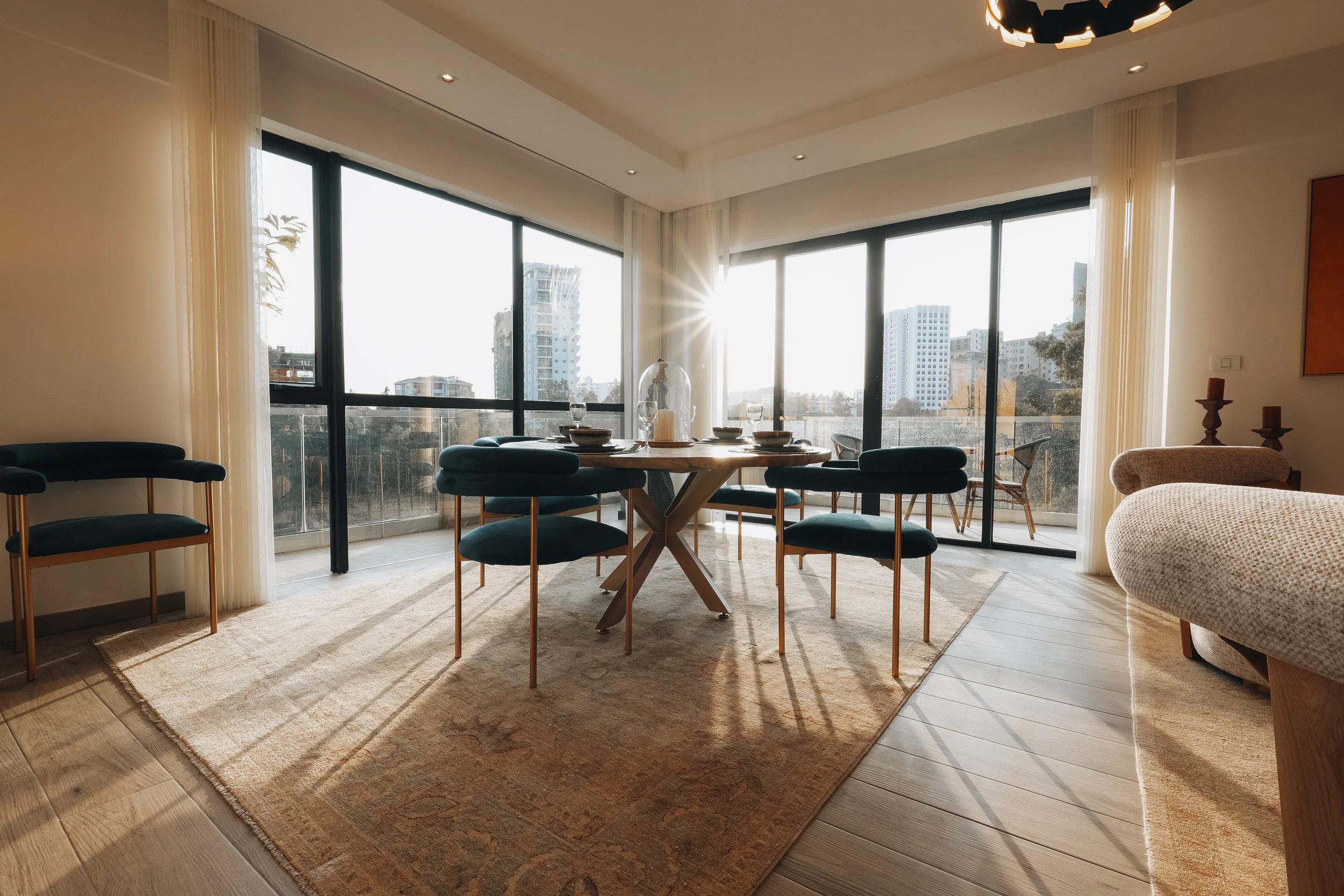 Modern dining room with large windows, round wooden table, six dark blue chairs, near balcony with outdoor furniture, sunlight streaming, beige rug, wooden floor, and minimal decor.