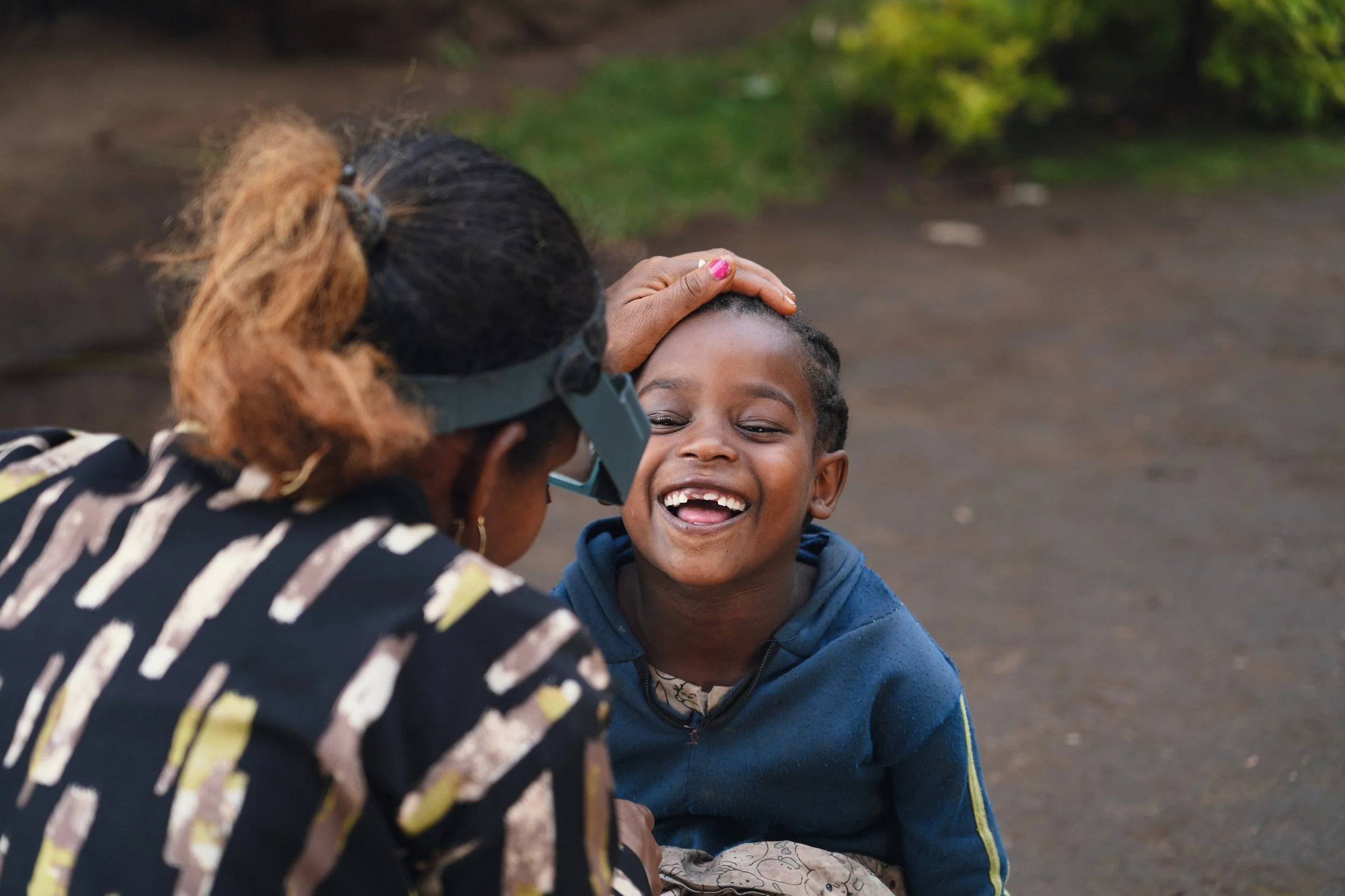 A woman wearing glasses and a striped jacket gently touching the head of a smiling young boy dressed in a blue hoodie.