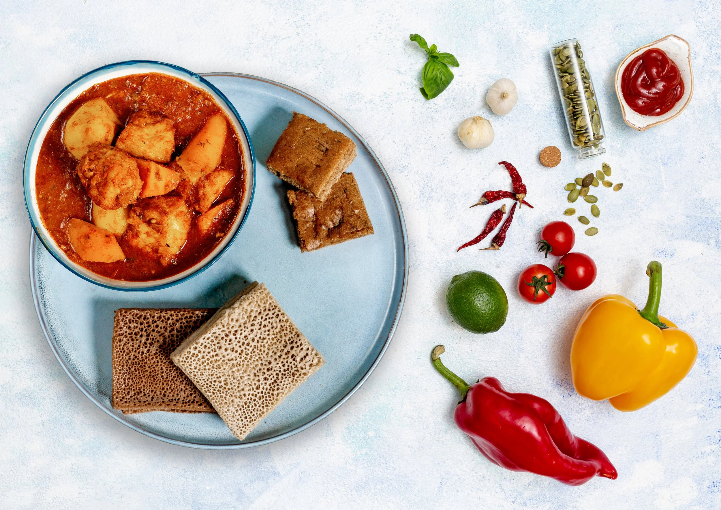 Indian food with a bowl of curry, papadums, and bread on a blue plate surrounded by tomatoes, lime, chili peppers, garlic, basil, pumpkin seeds, and condiments.