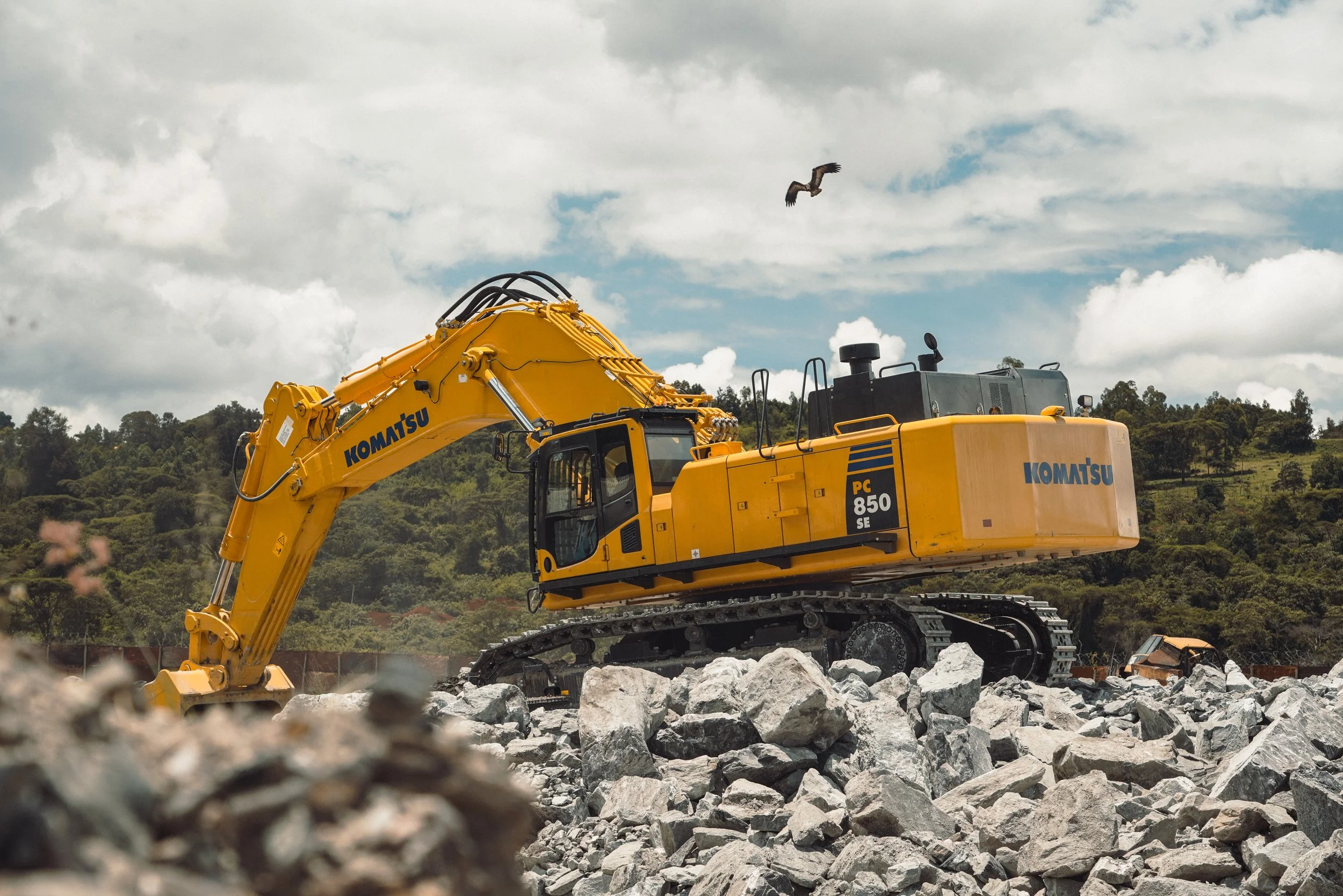 A yellow Komatsu excavator working on a rocky construction site with a hill and partly cloudy sky in the background.