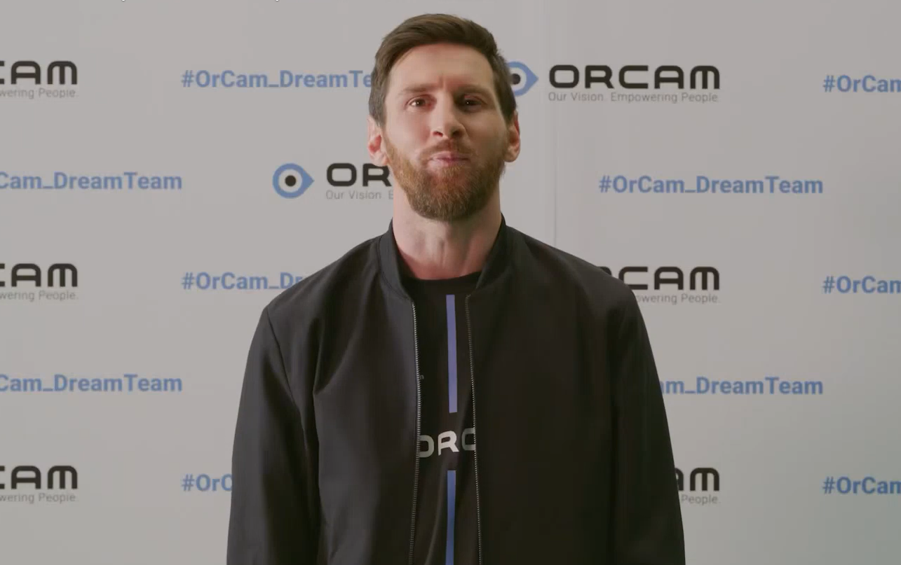 A man standing in front of a backdrop with the ORCAM logo and hashtags, wearing a black jacket and a black shirt with the company's name.
