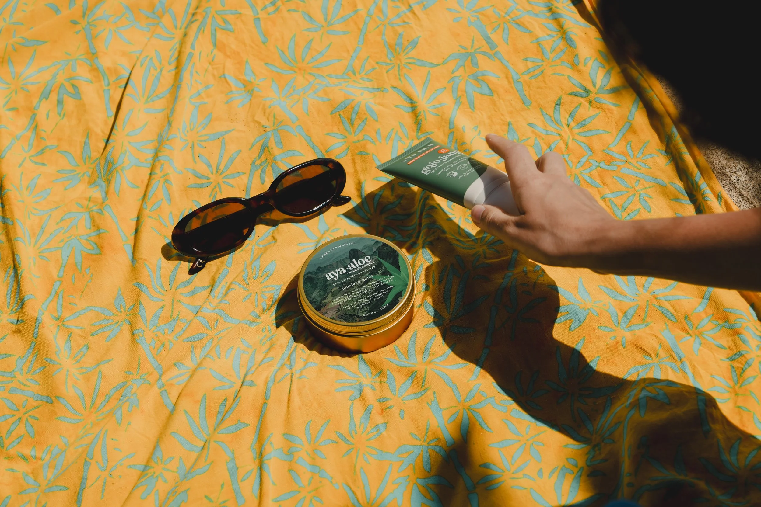 A pair of sunglasses, a container of aya-aloa skincare product, and a tube of sunscreen are placed on a yellow fabric with a blue leaf pattern. A person's hand is reaching towards the sunscreen.