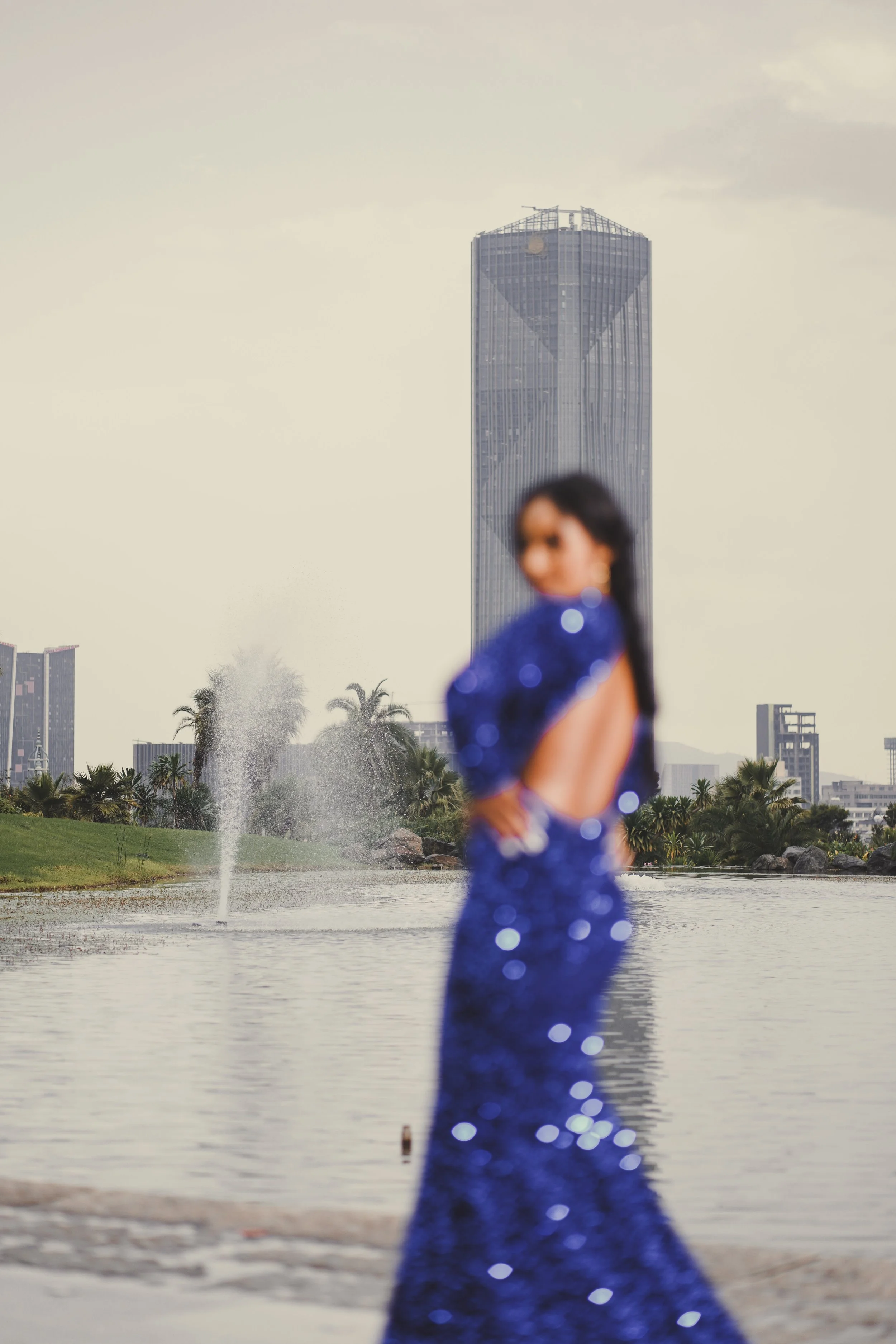Blurry woman in a shiny blue dress standing near a body of water with a city skyline, including a tall hexagonal skyscraper and palm trees, in the background.