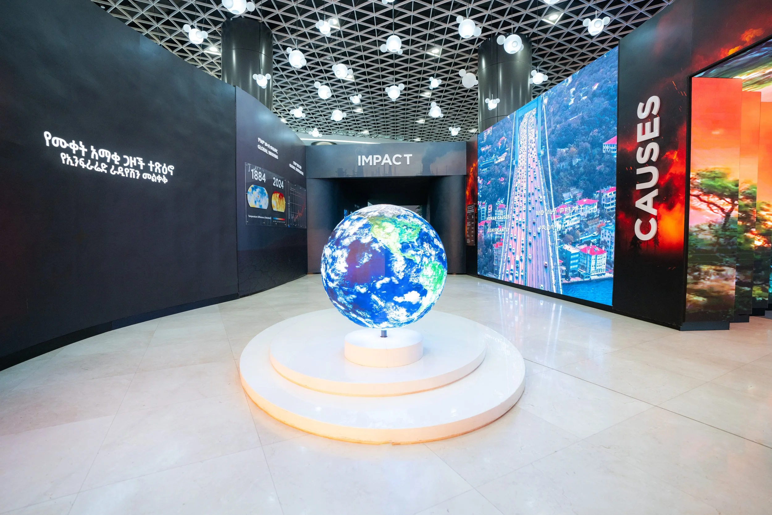 Exhibit with a globe displaying Earth on a white platform, surrounded by informational displays and large digital screens showing images of cityscapes and pollution, with the words 'IMPACT' and 'CAUSES' visible.