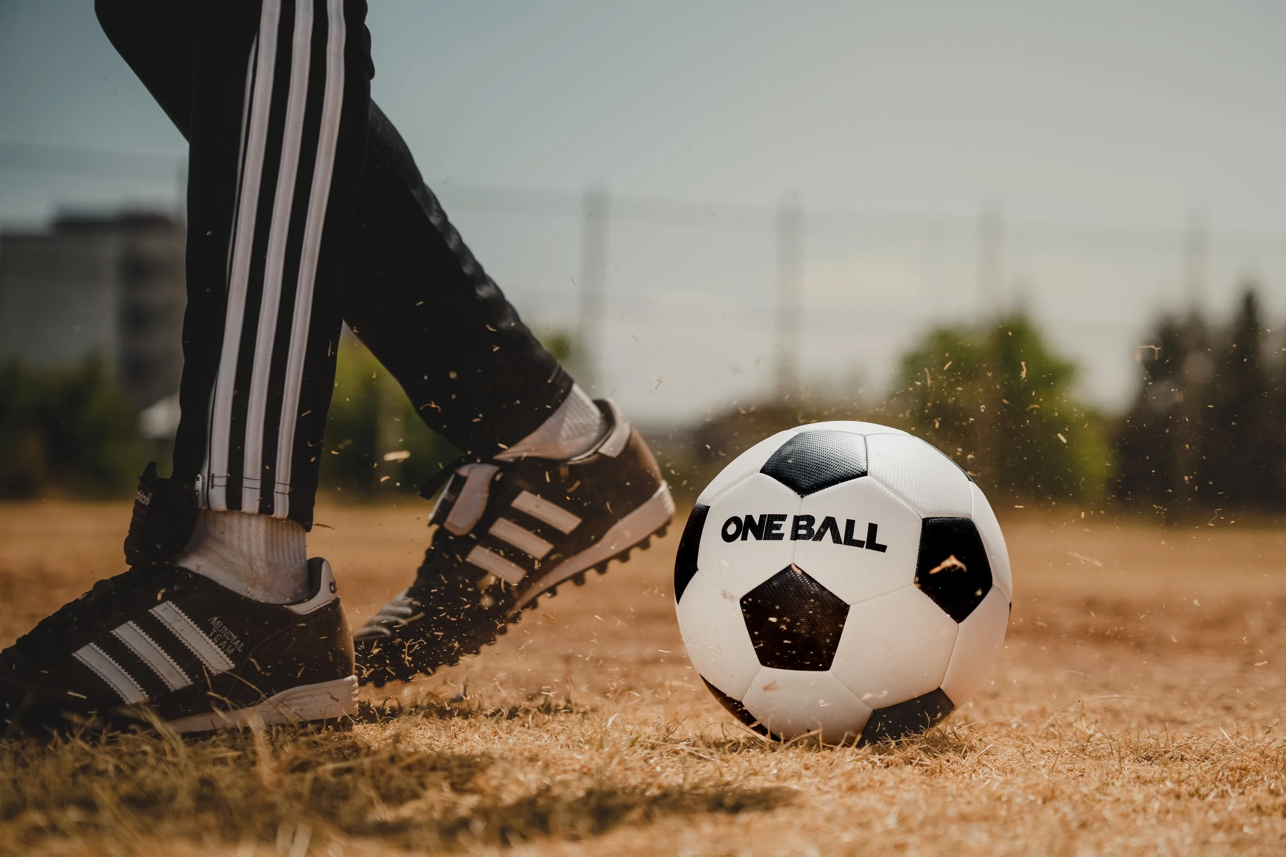 A person wearing black Adidas sneakers with white stripes and white socks is preparing to kick a white and black soccer ball on a dirt field. The soccer ball has the words "ONE BALL" written on it.