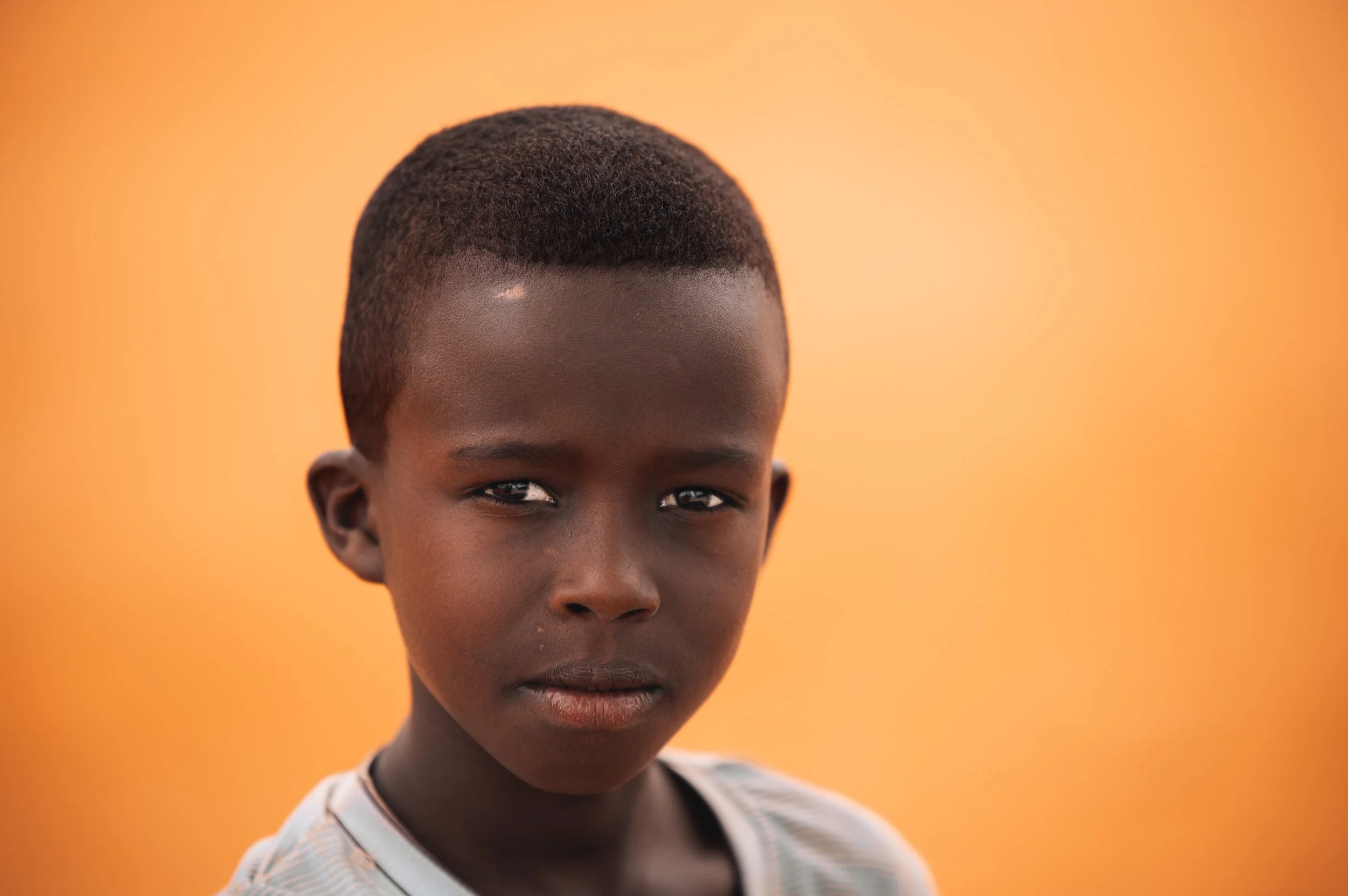 Portrait of a young boy with dark skin and short hair against an orange background.