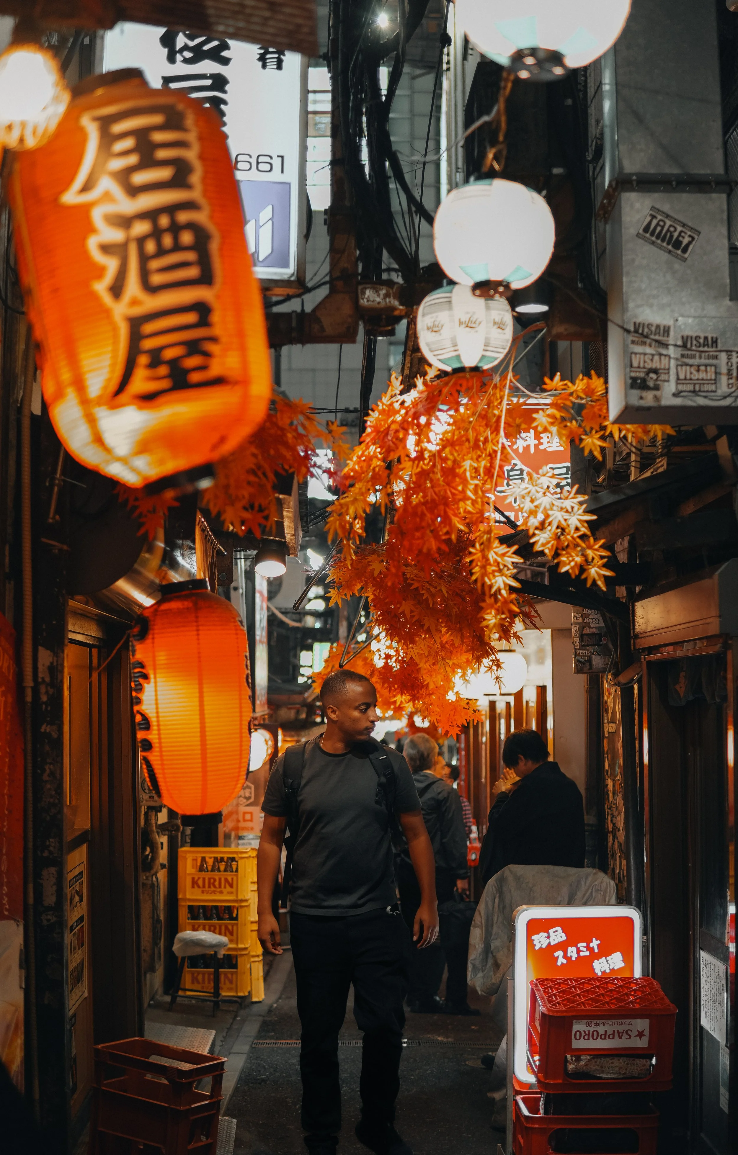 A narrow street scene at night in Japan, illuminated by orange and white lanterns hanging overhead, with autumn leaves decorations. A man is walking through the street, with people in the background near shops and restaurant entrances.