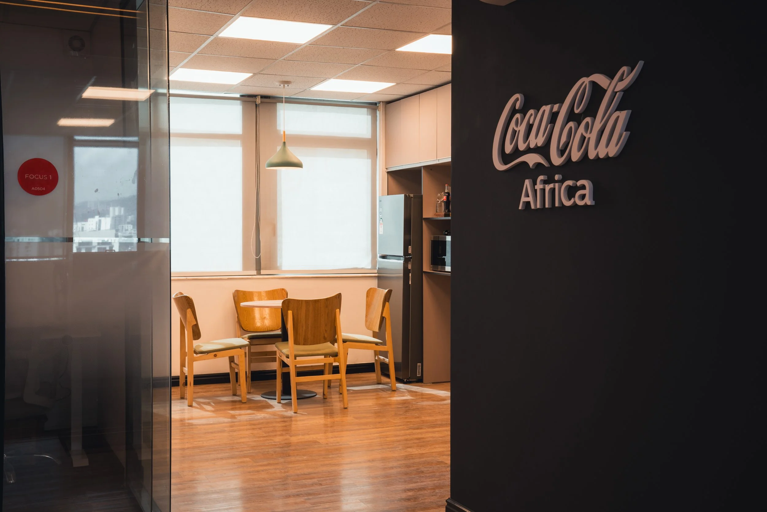 Interior of an office with wooden flooring, a small round table with four wooden chairs, large windows with natural light, and a sign on a dark wall that reads 'CocaCola Africa'.