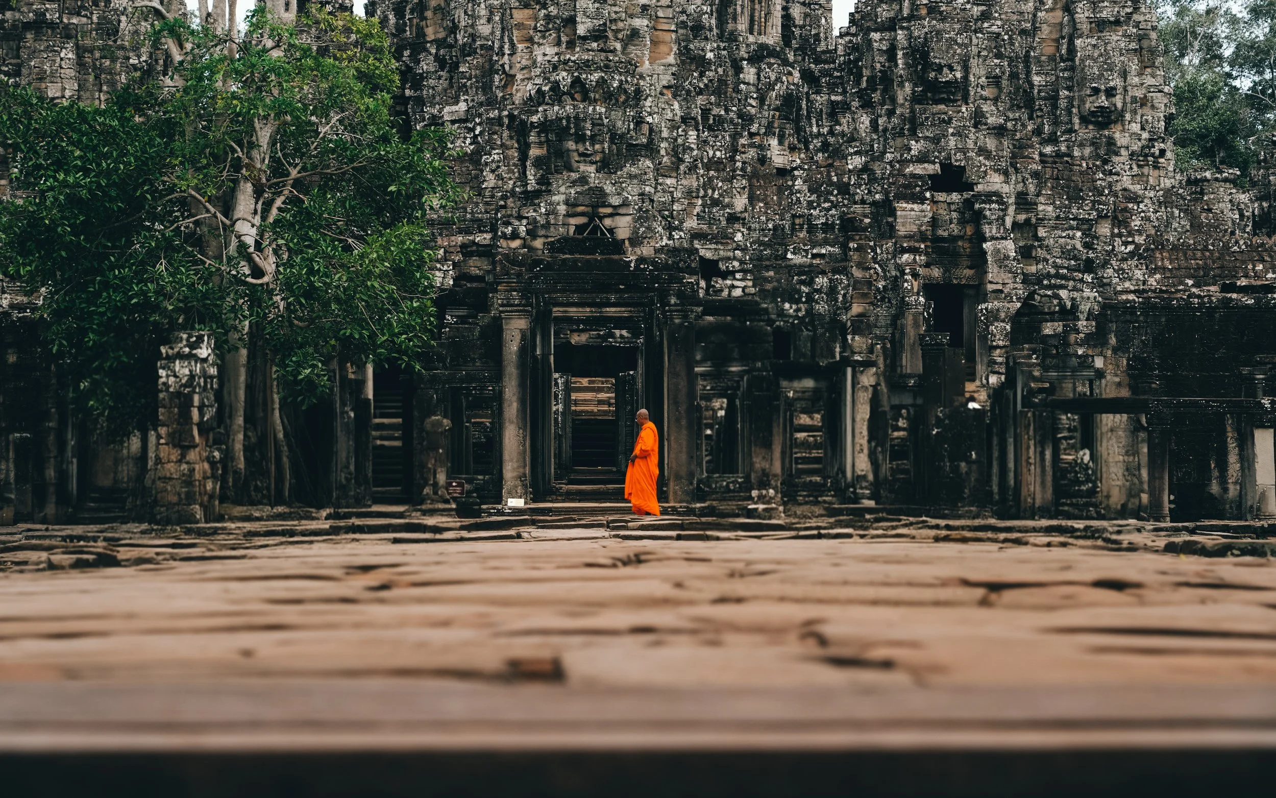 A Buddhist monk in an orange robe standing in front of an ancient stone temple surrounded by trees.