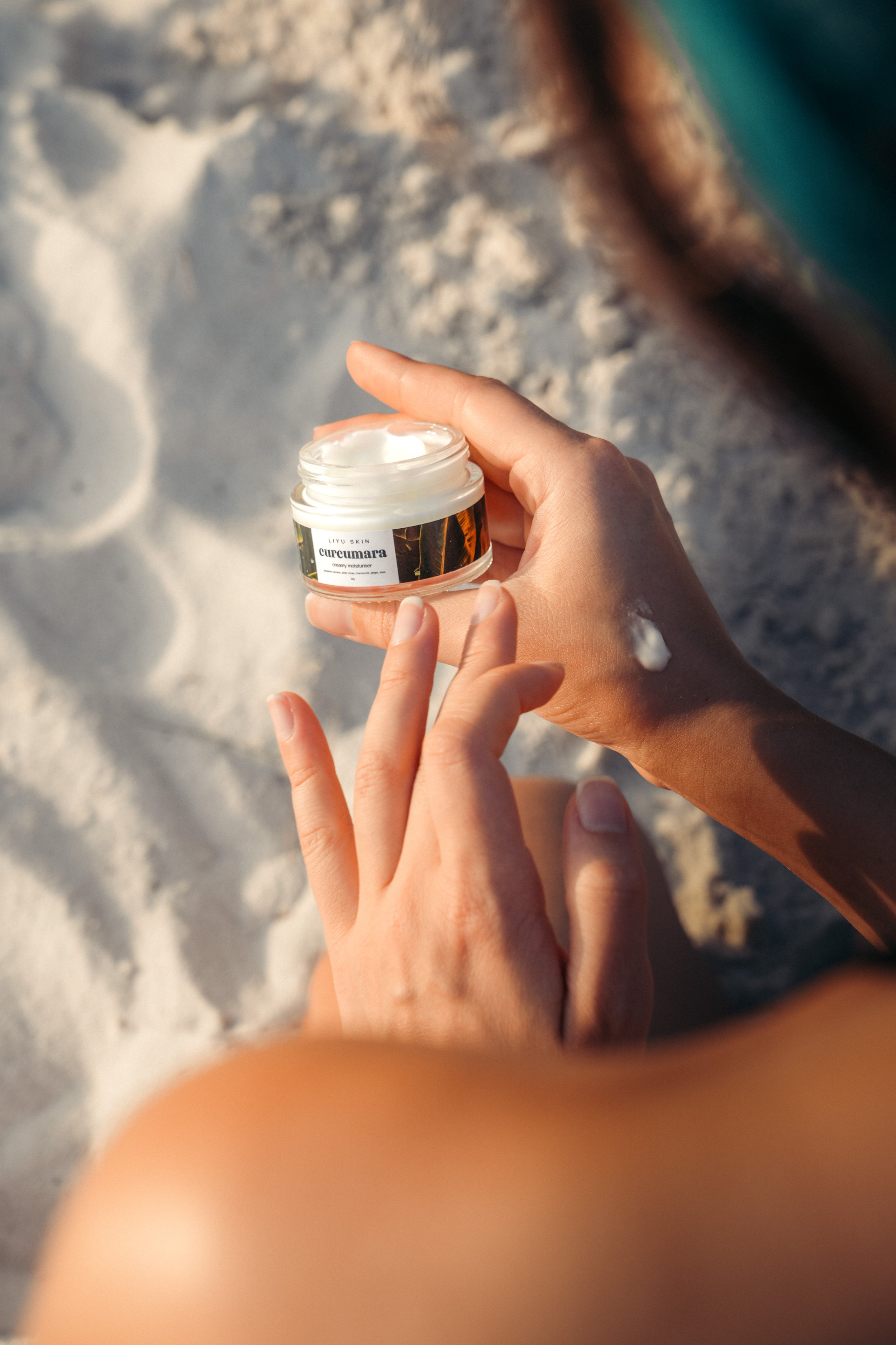 A person holding an open jar of cream on a sandy beach with a bit of lotion on their hand.