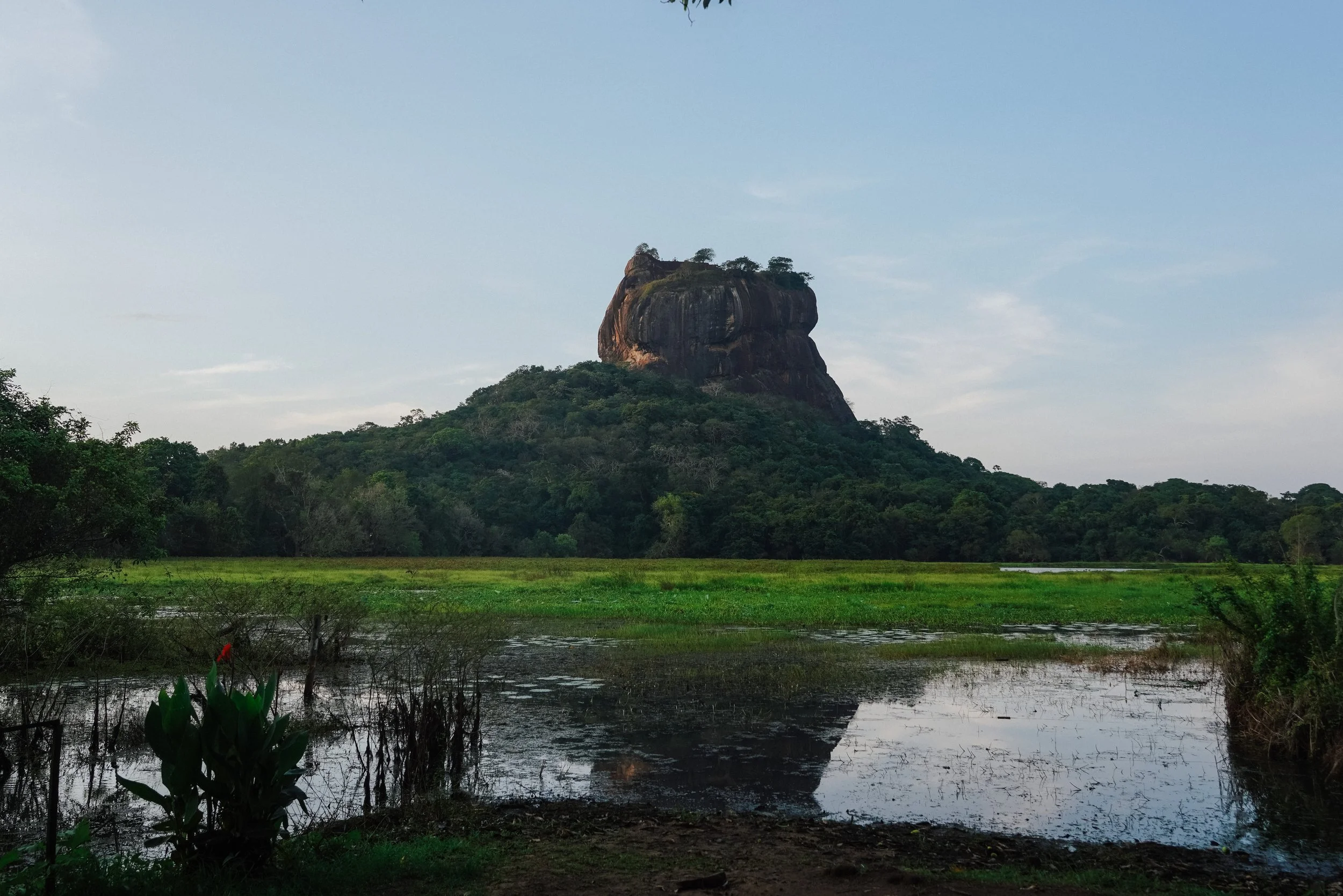A large rocky hill or mountain with a flat top, surrounded by lush green trees and vegetation, with a body of water in the foreground reflecting the hill.