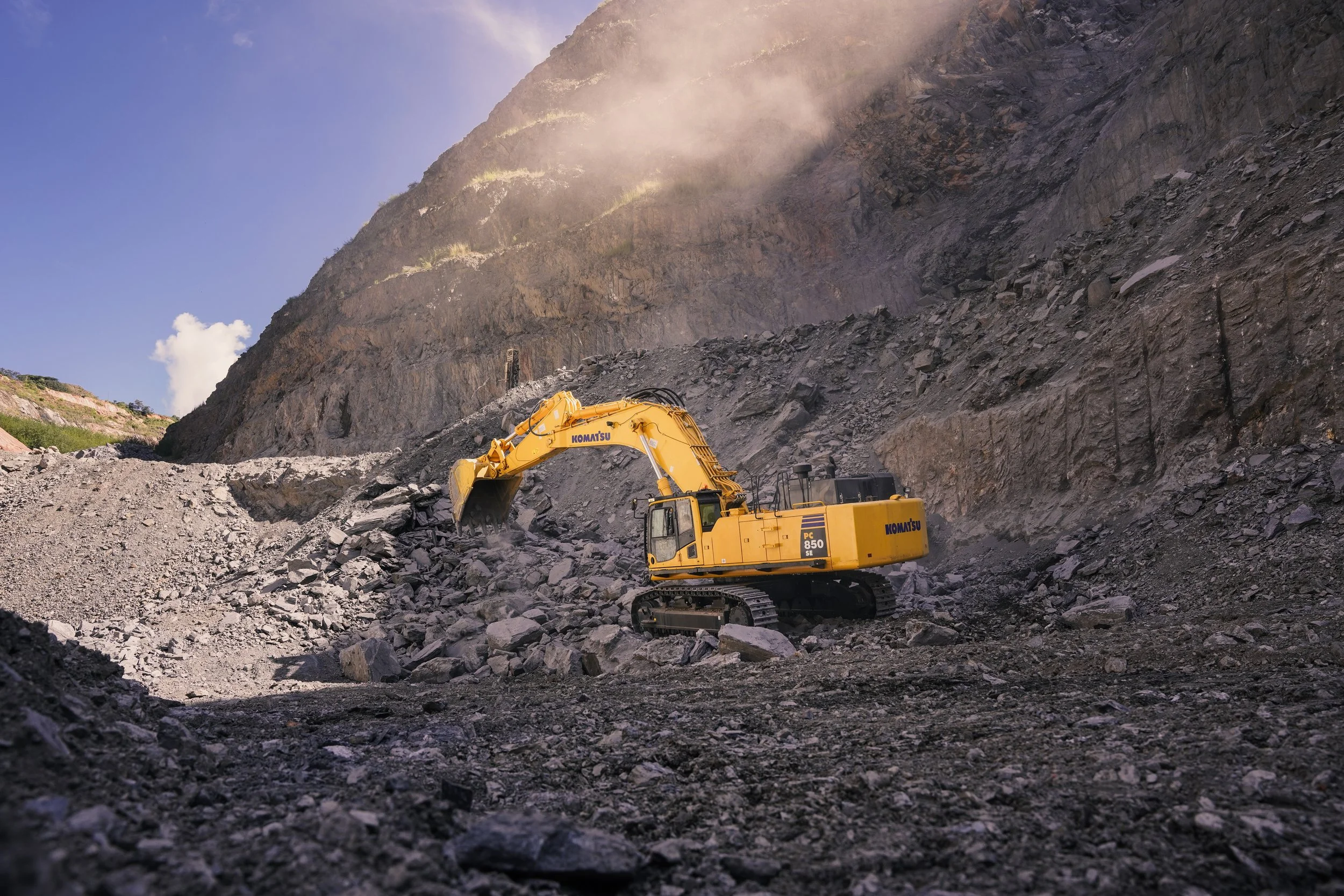 A yellow Komatsu excavator working in a rocky mountainous terrain with a steep slope in the background.