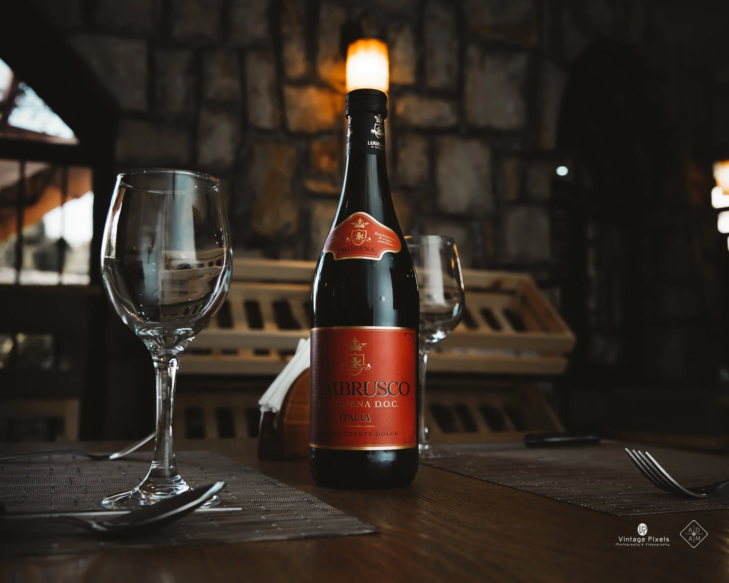 A bottle of Lambrusco wine on a wooden table, flanked by two empty wine glasses, in a rustic restaurant setting with stone walls and warm lighting