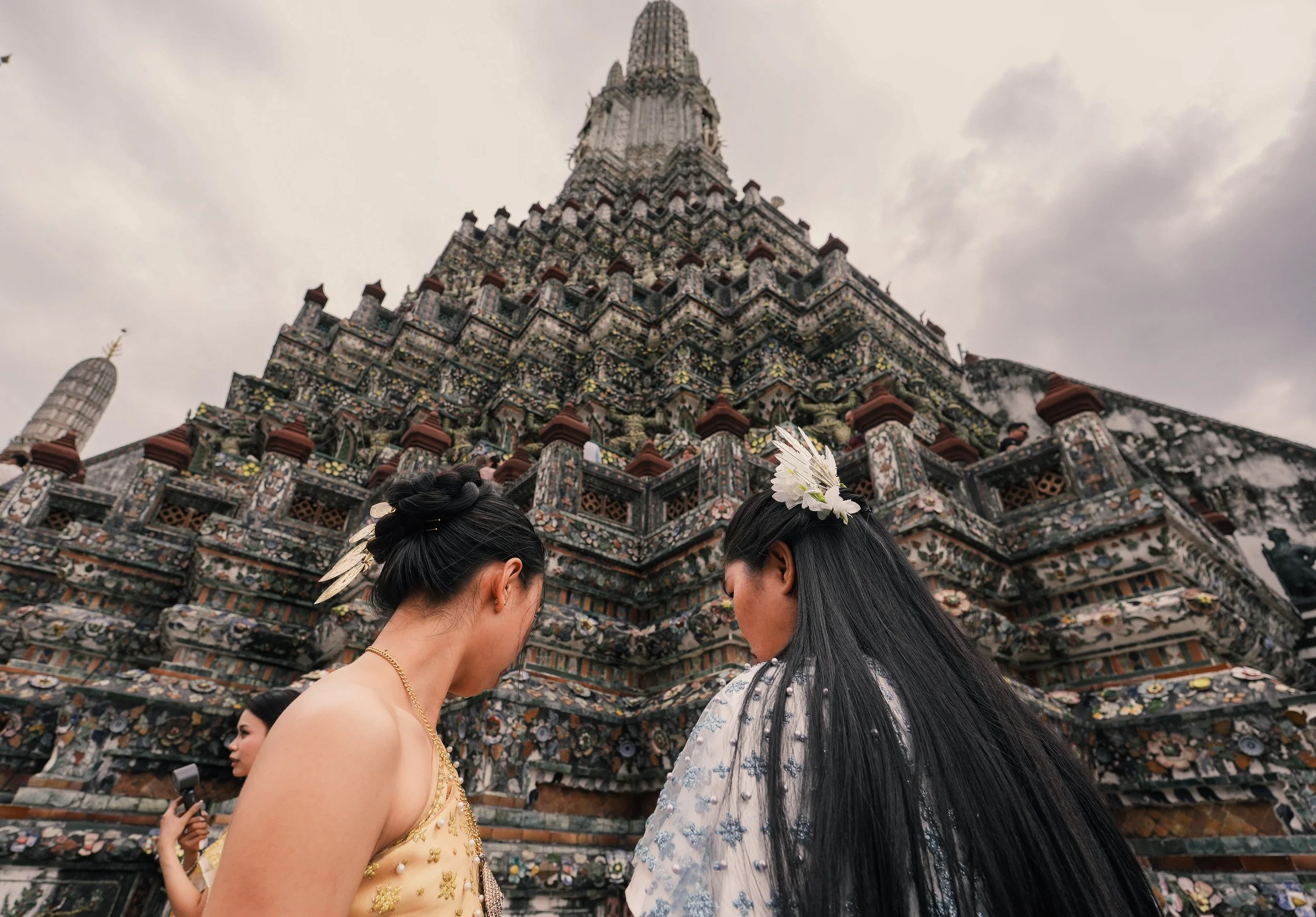 Two women with long dark hair and flower hair accessories praying in front of an ornate temple decorated with colorful mosaics.