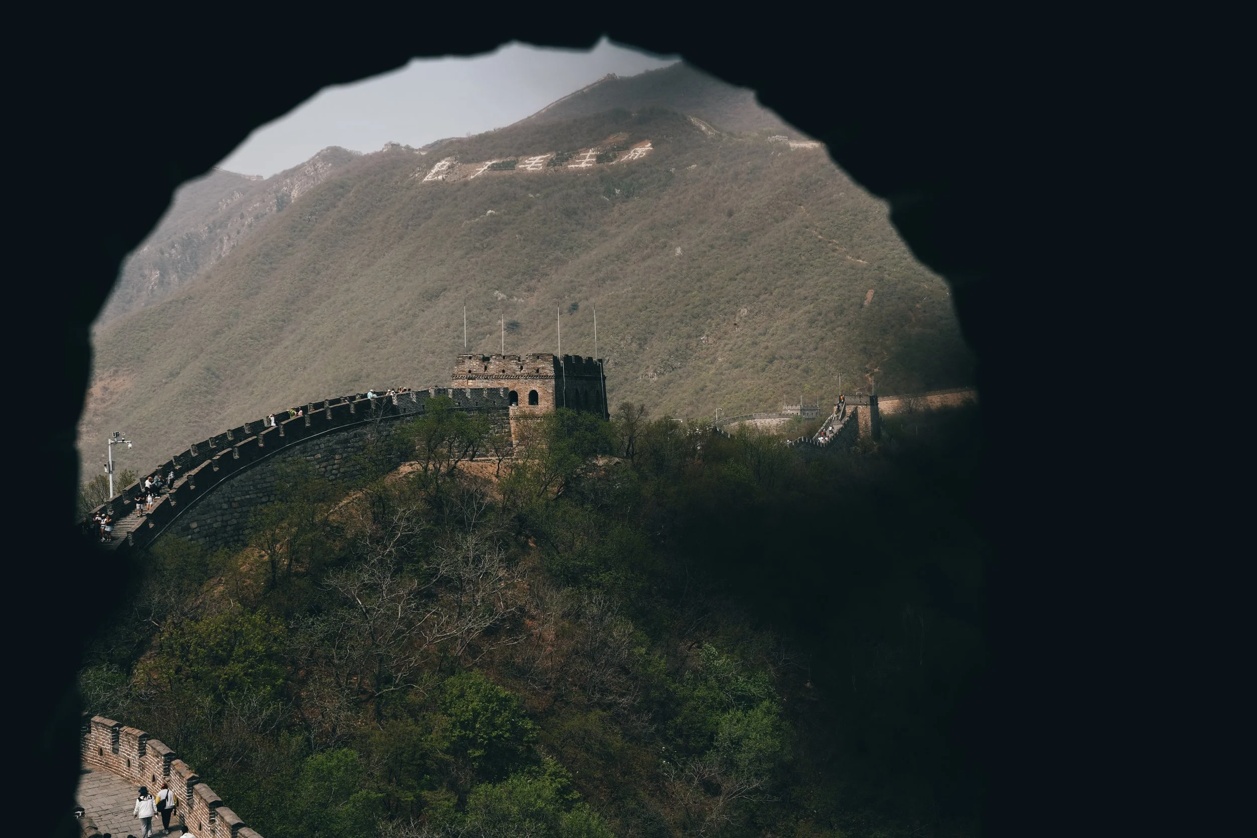 The Great Wall of China viewed through a stone or brick archway, with lush green hills and mountains in the background.