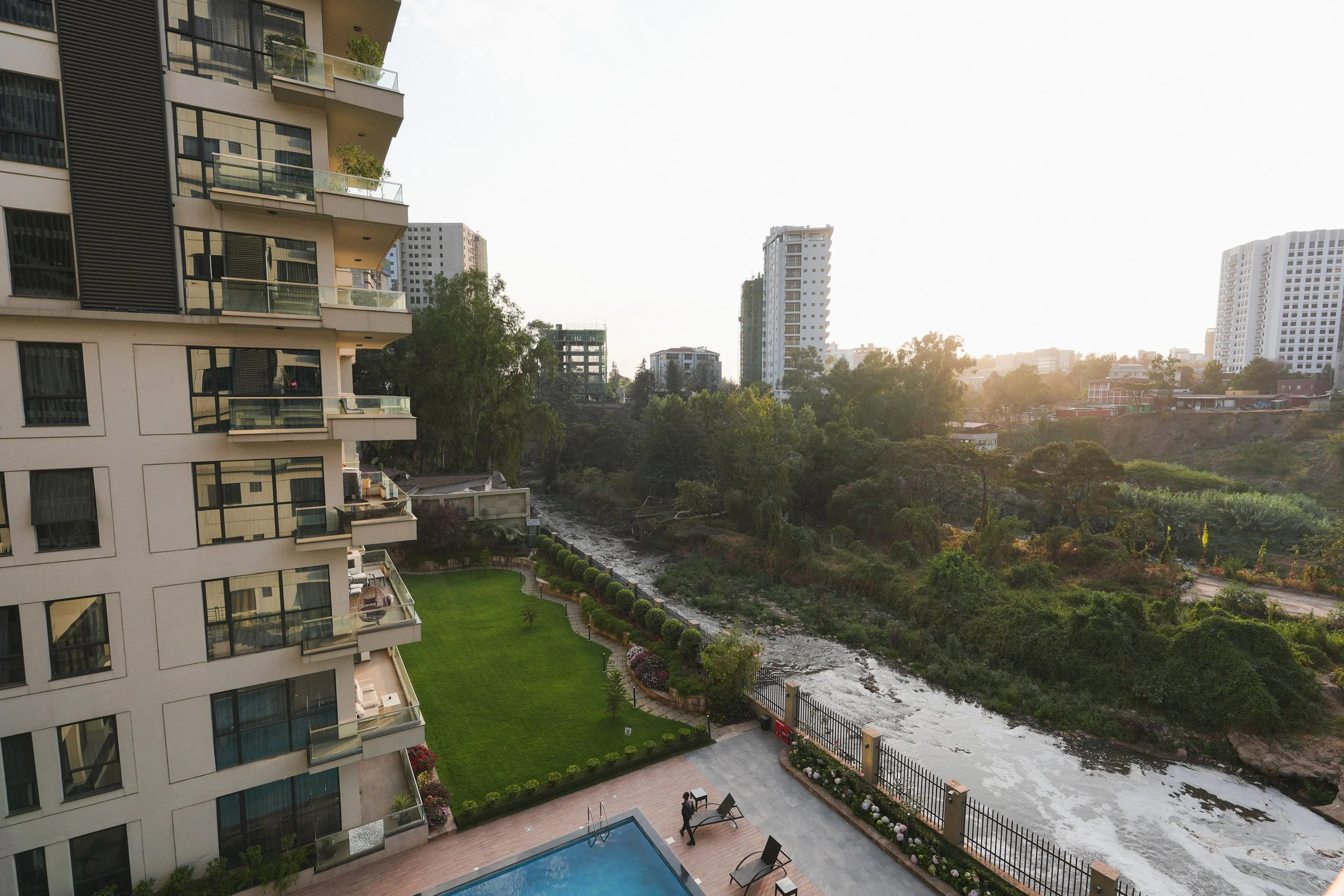 Urban residential area with a modern apartment building, a swimming pool, a lawn, and a creek with trees and other buildings in the background during sunset.