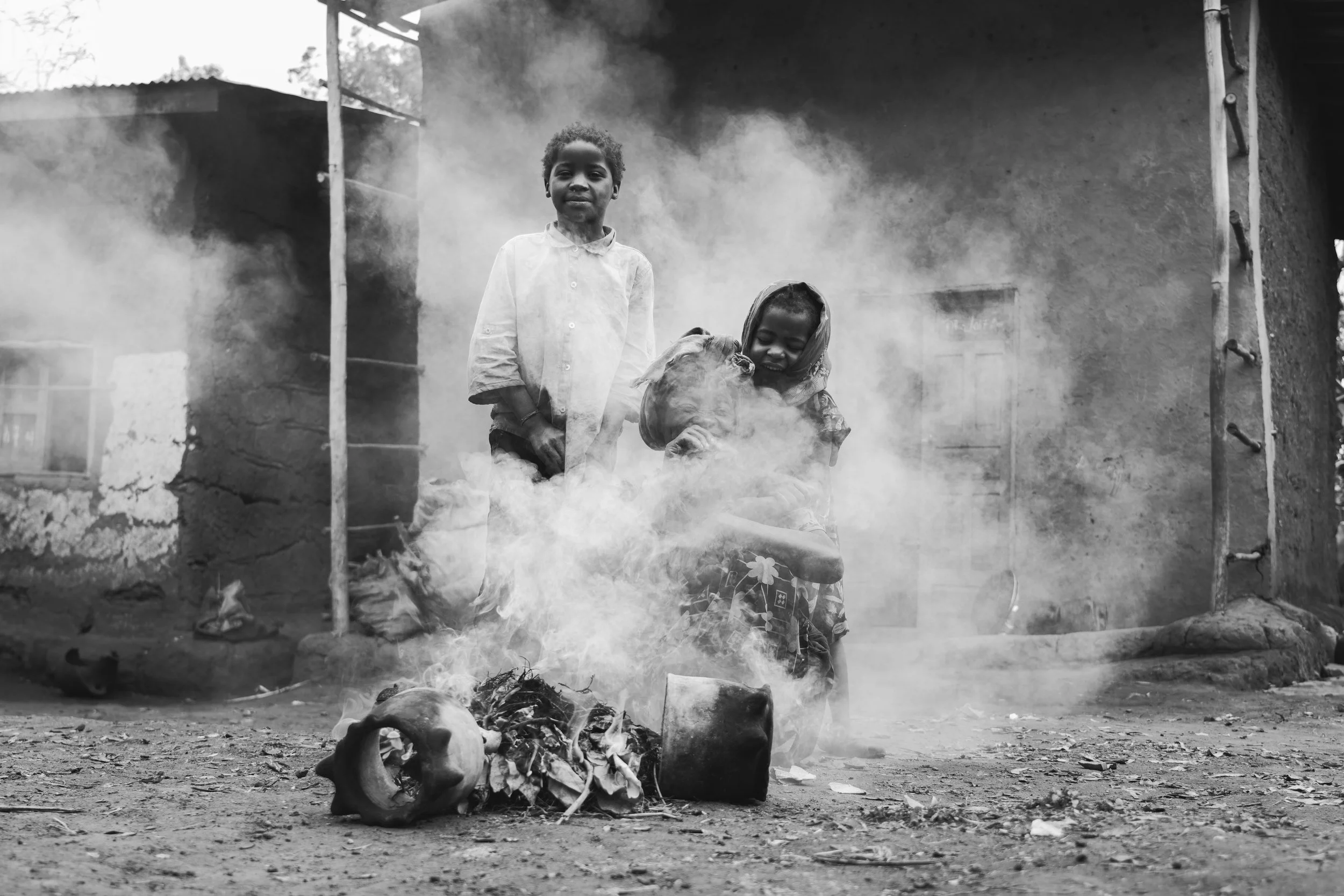Two children, a boy and a girl, stand near a small fire with smoke, outside a rustic building made of mud or clay, in a rural setting.