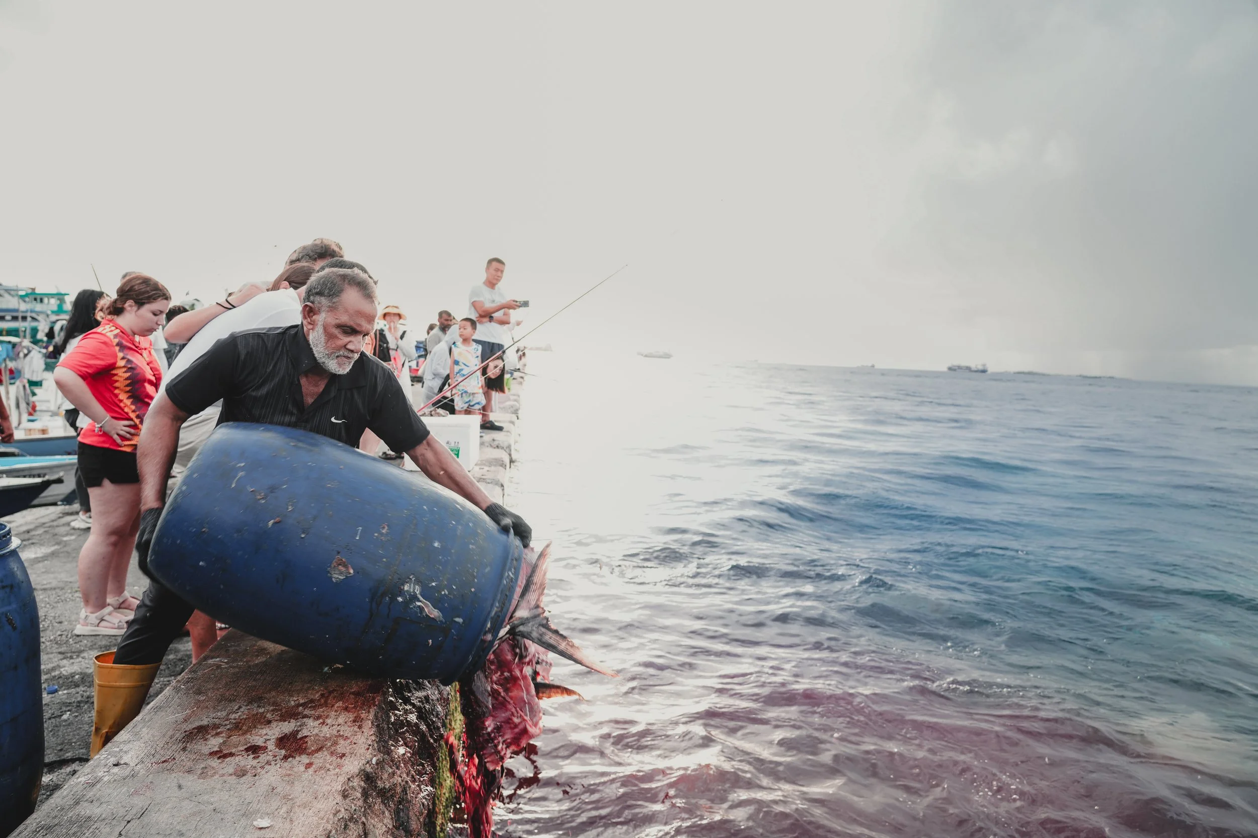 A man in black clothes and yellow boots is pouring water from a blue barrel into the sea, surrounded by people on a dock, with boats in the background on a cloudy day.