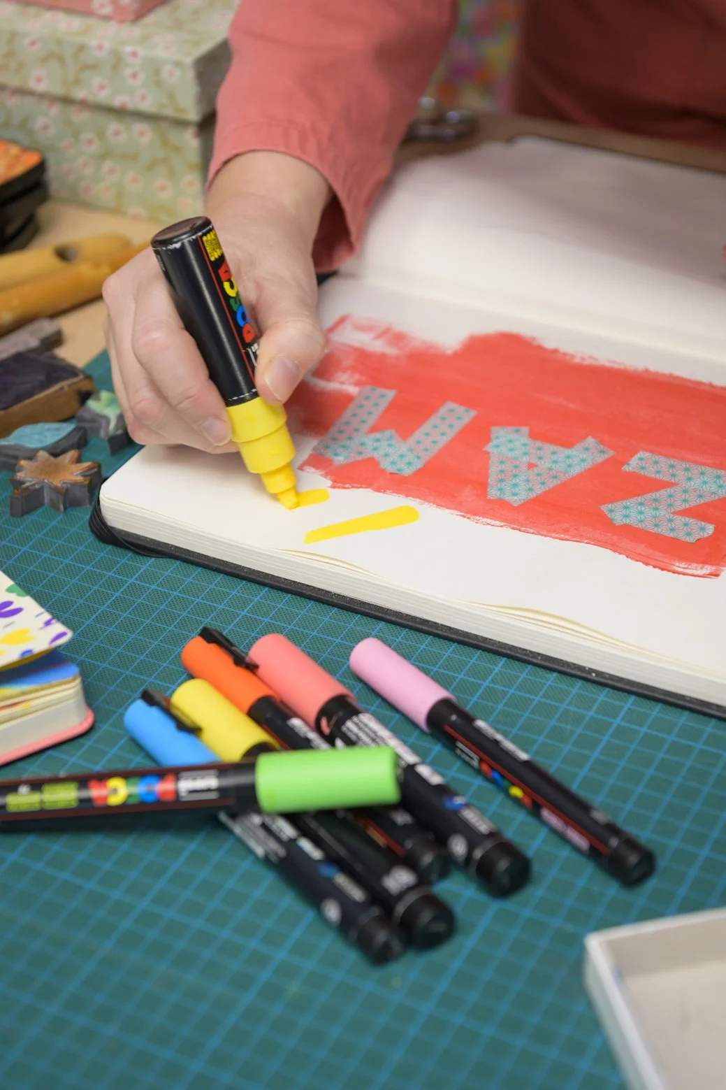 Une personne utilise un marqueur jaune pour colorier une page dans un carnet, créant une graphie avec des lettres en couleurs et motifs sur une surface de travail avec divers marqueurs et fournitures d'art.