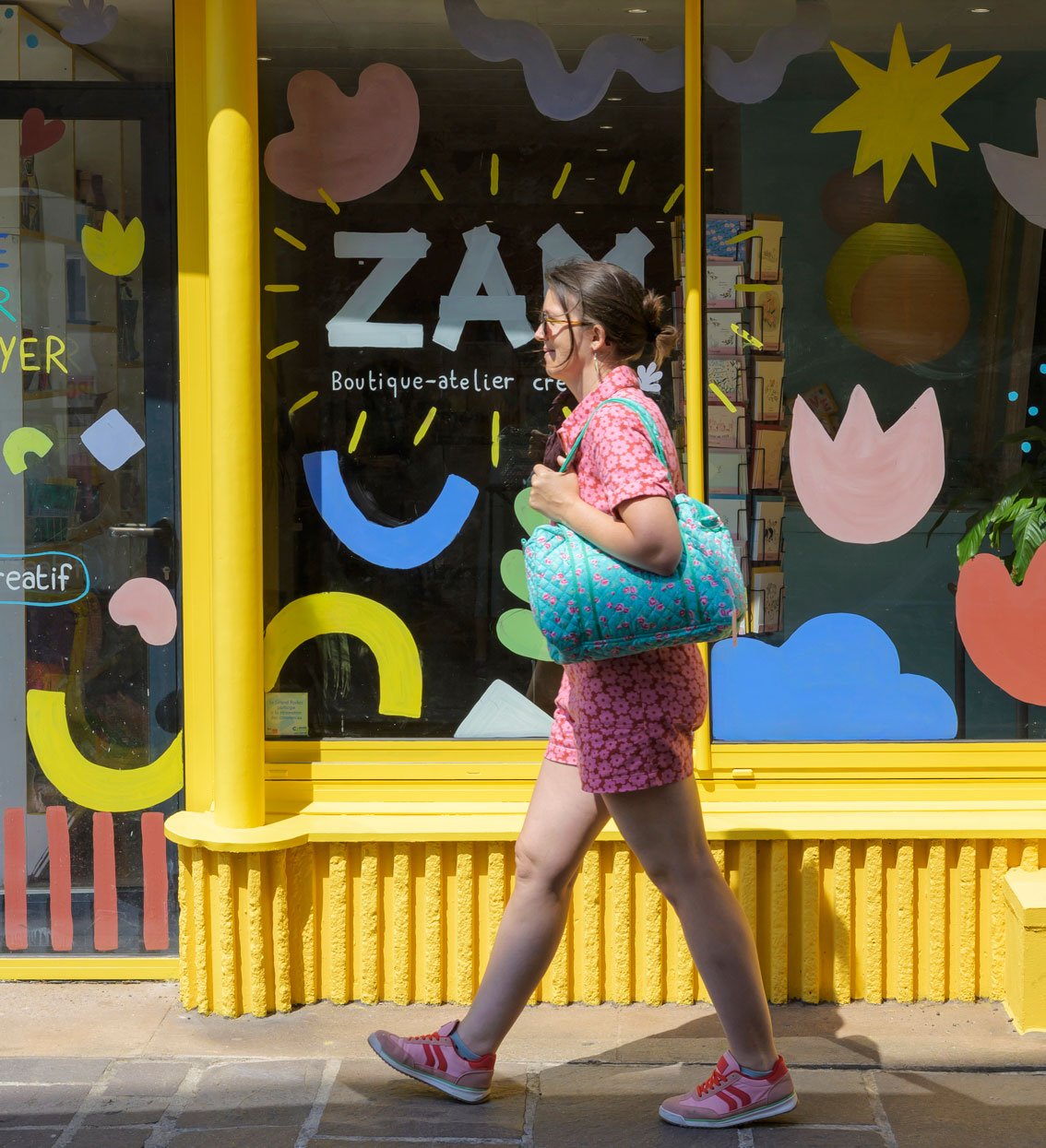 Femme marchant devant un magasin de boutique créative avec vitrine colorée, portant une robe rose avec motifs floraux et un sac à dos turquoise, ensoleillée.