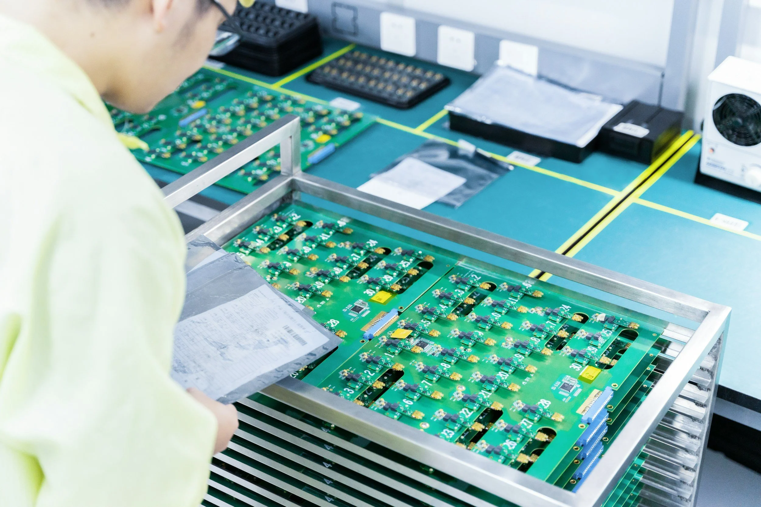 Technician inspecting green circuit boards with electronic components on a worktable in a manufacturing or repair facility.