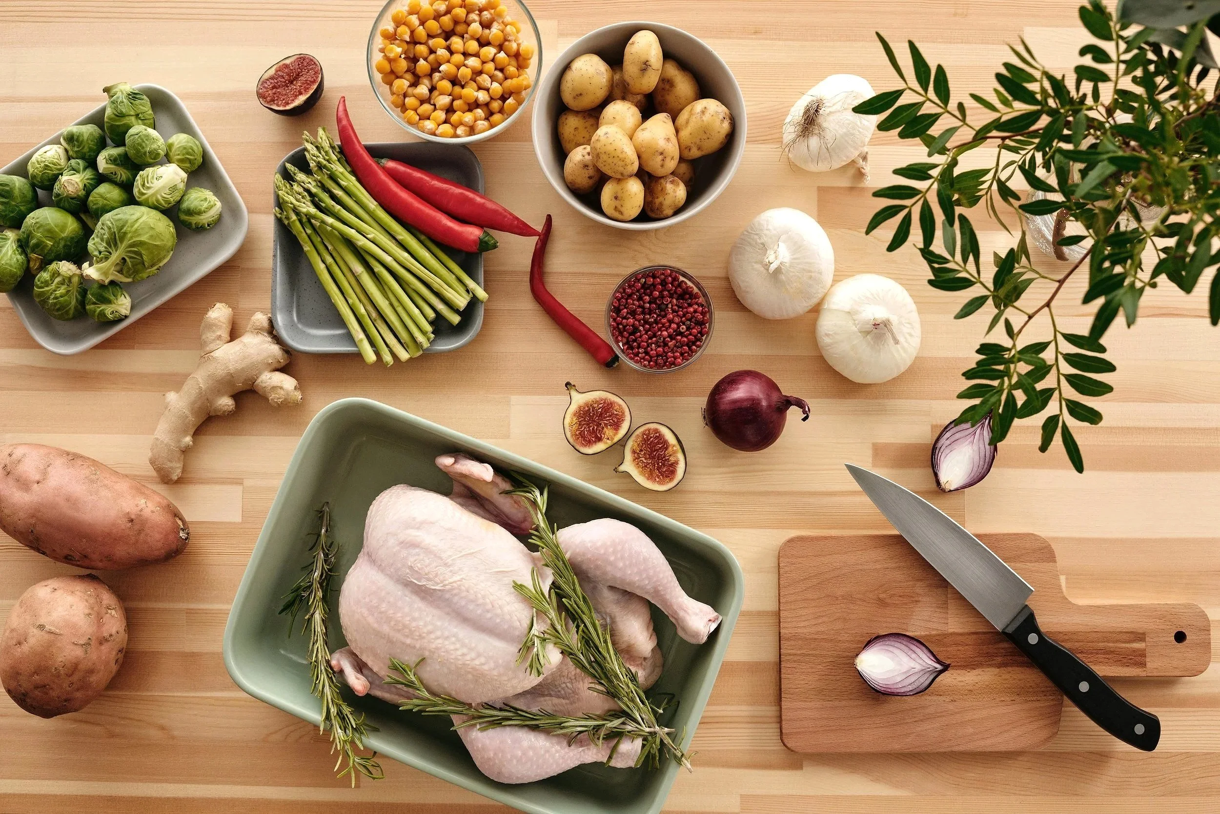 A wooden kitchen countertop with fresh vegetables, spices, and a whole raw chicken garnished with rosemary, next to a cutting board with a knife and garlic cloves, along with potatoes, brussels sprouts, asparagus, red chilies, garlic, onions, figs, corn, and various herbs.