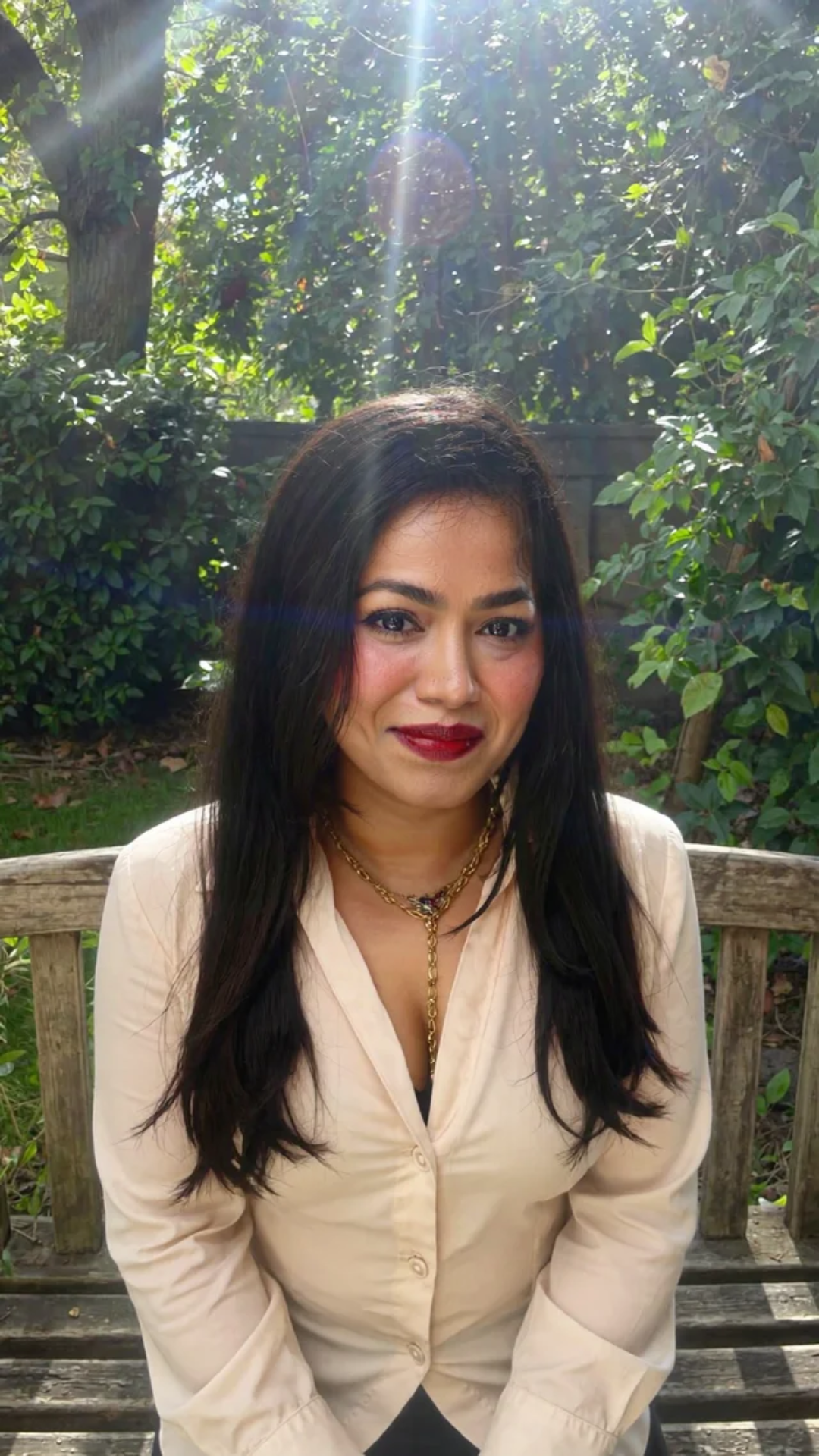 A woman with long dark hair, red lipstick, wearing a beige blouse and layered gold necklaces, sitting on a wooden bench outdoors with lush green foliage and sunlight in the background.