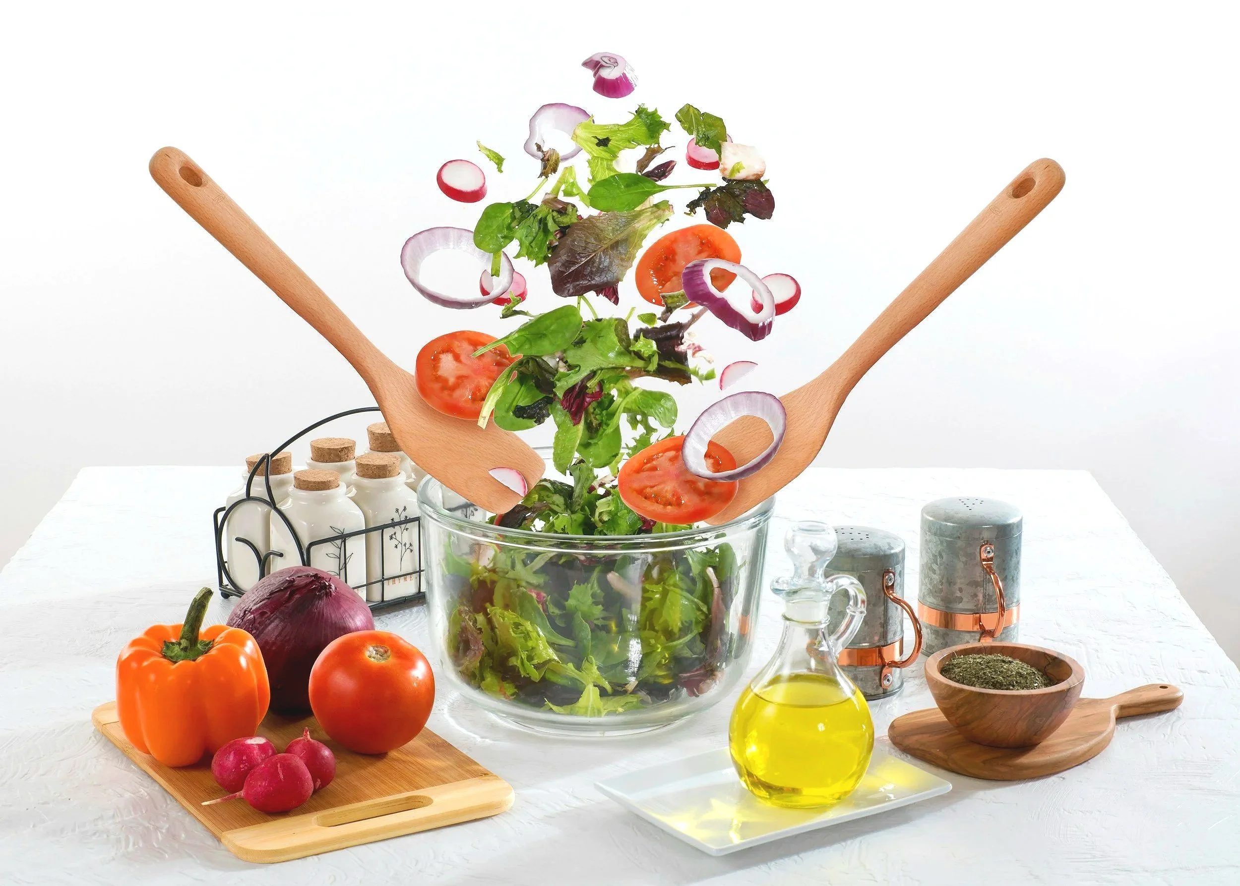 Salad ingredients, including cherry tomatoes, radishes, red onion, and mixed greens, are being tossed in a glass bowl with two wooden salad servers. Whole vegetables such as yellow pepper, red onion, and tomatoes are on a wooden cutting board, along with a small bunch of radishes. A small bottle of olive oil, salt and pepper shakers, and a bowl of herbs are also on the white table.