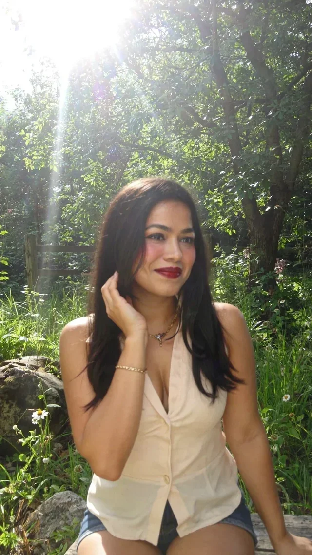 A young woman with long dark hair, wearing a beige sleeveless top and denim shorts, sitting outdoors in a lush green setting with sunlight filtering through the trees.