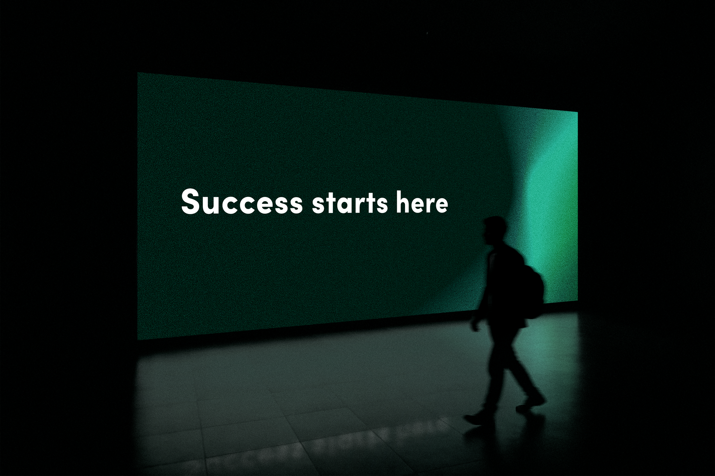 A digital sign with the words "Success starts here" displayed in white letters on a dark green background, with a silhouette of a person walking in front of it in a dark, indoor setting.