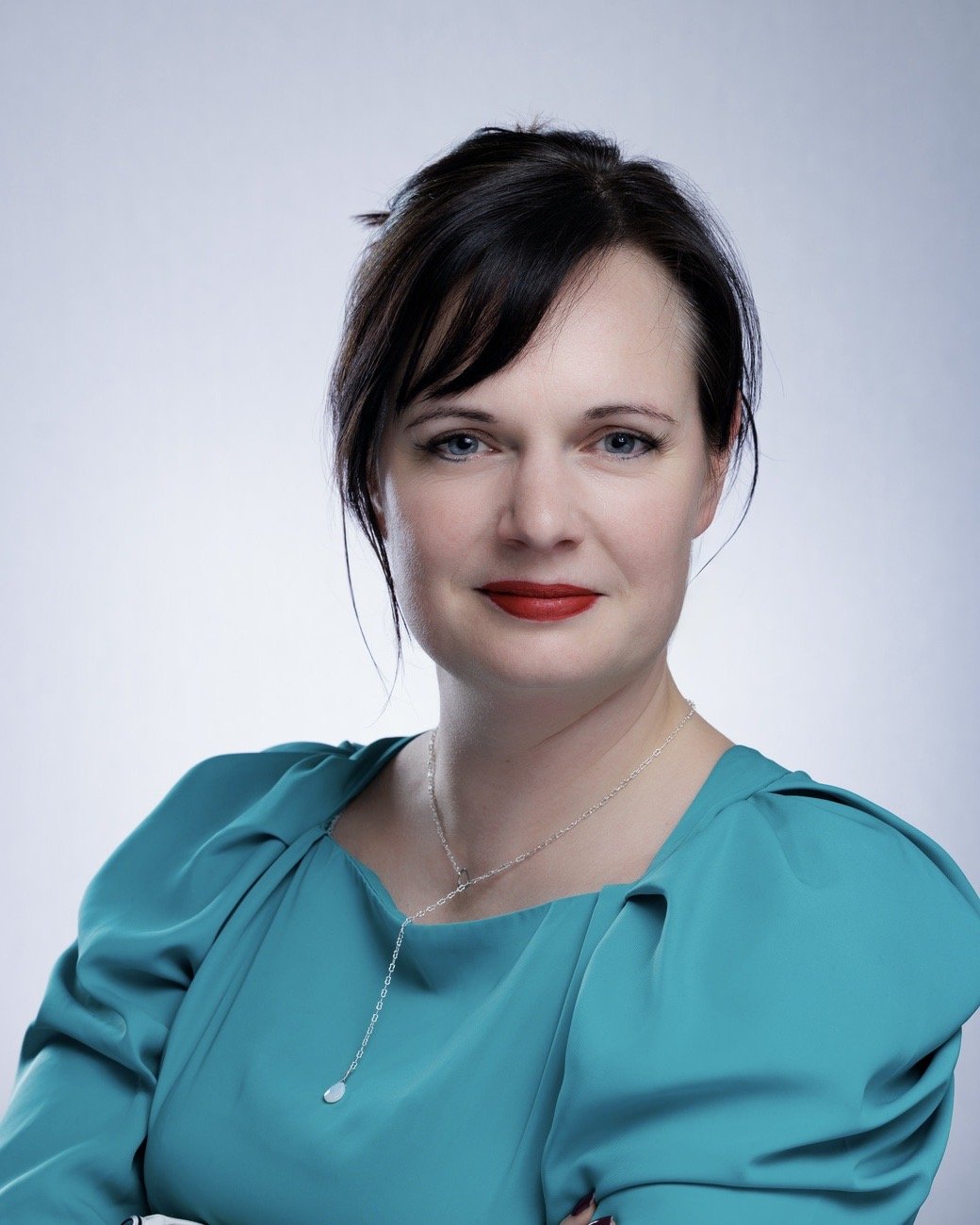 A woman with dark brown hair in a short haircut, wearing a teal dress with puffed sleeves, red lipstick, and a delicate silver necklace, posing against a plain light gray background.