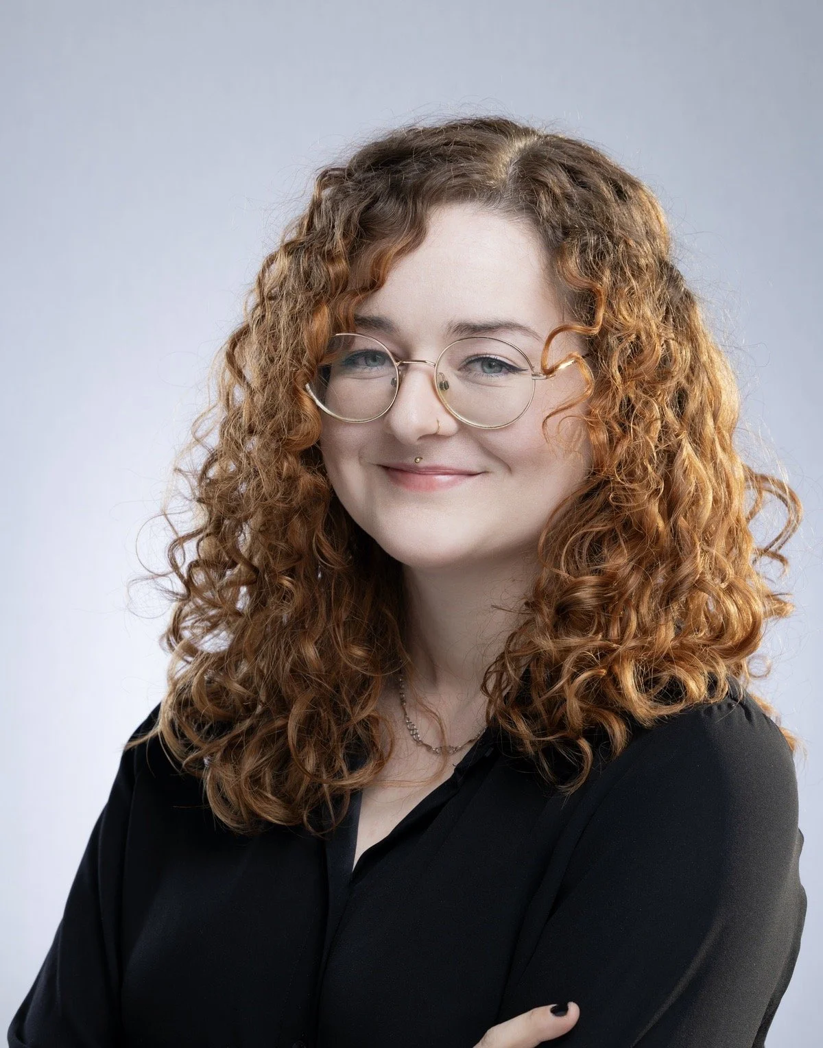 A young woman with curly red hair wearing glasses and a black top, smiling at the camera against a plain background.