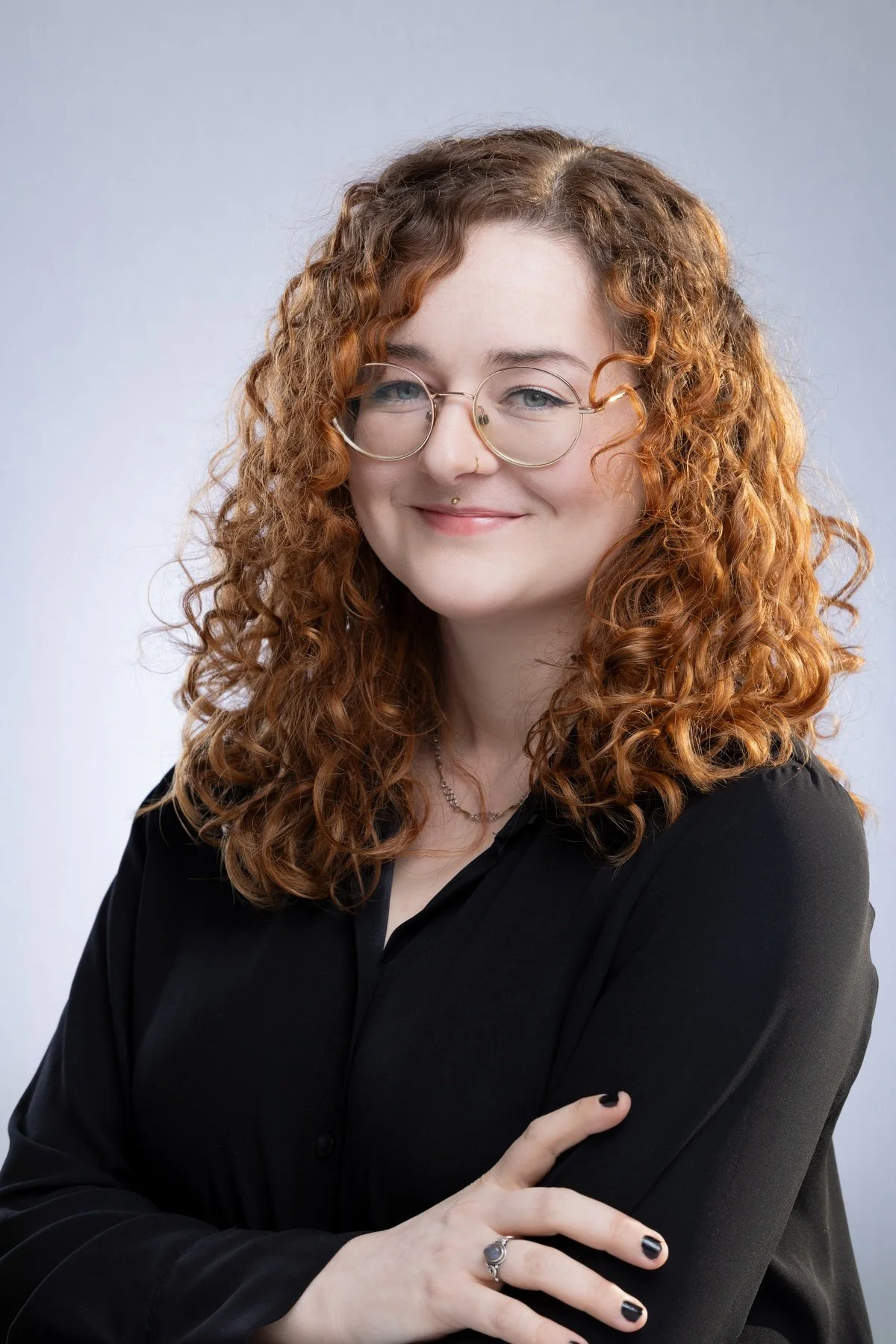 A woman with curly red hair, wearing glasses, a nose ring, and a black top, smiling in front of a plain gray background.