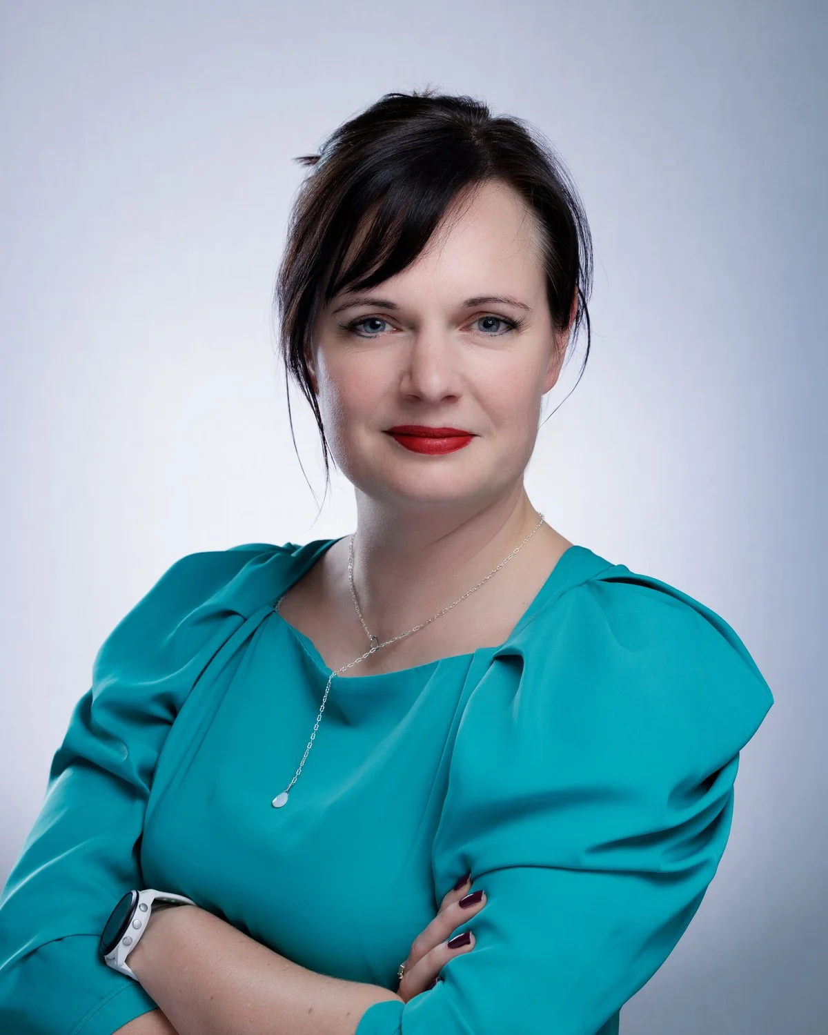 Portrait of a Hannah Vickers with short dark hair, blue eyes, red lipstick, wearing a teal blouse and a silver necklace, posed with arms crossed against a plain background.