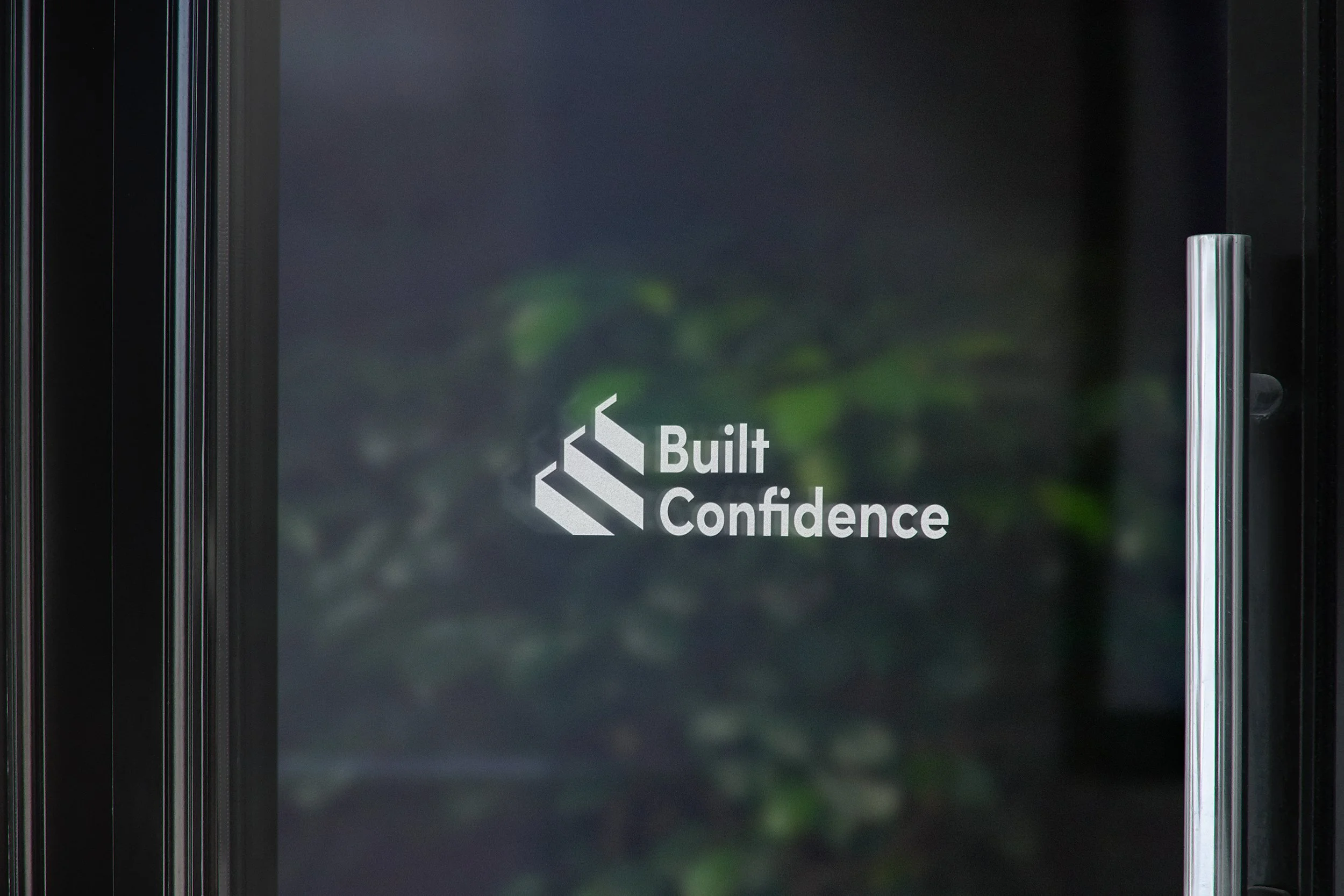 A glass door with a logo that says 'Built Confidence' and a staircase icon, with a blurred background of green plants.