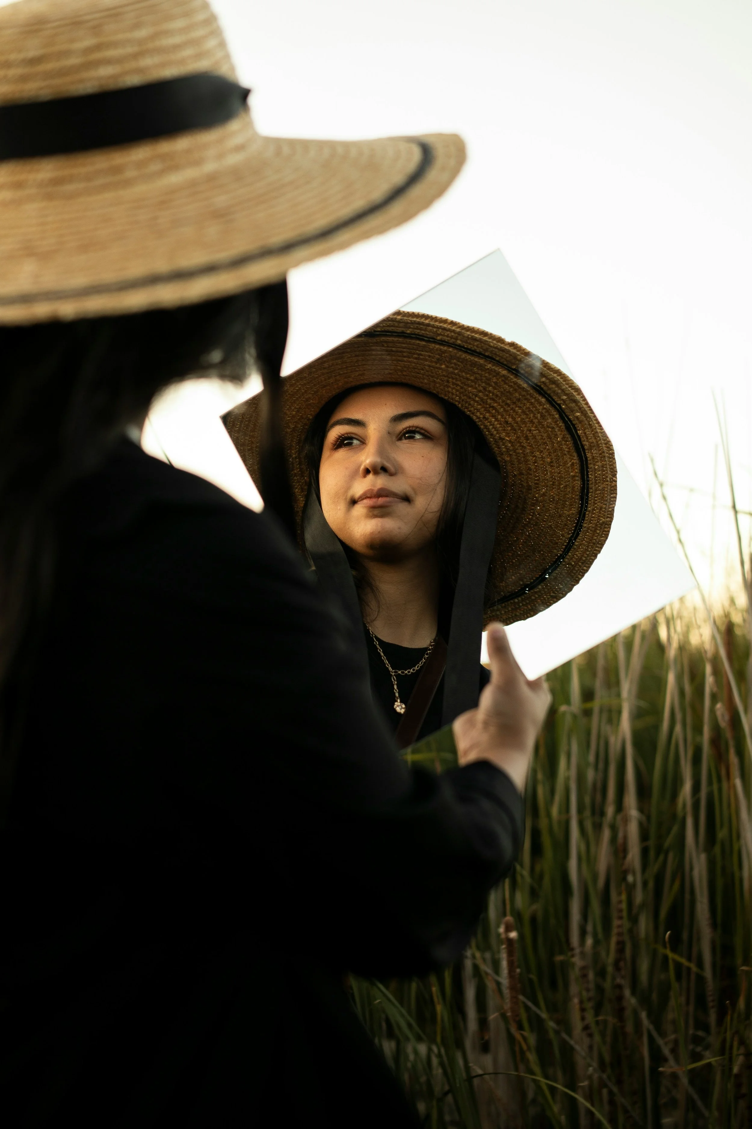 A woman in a straw hat looking into a handheld mirror, with tall grass in the background.
