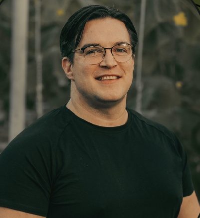 A young man with dark hair, glasses, and a black t-shirt smiling outdoors
