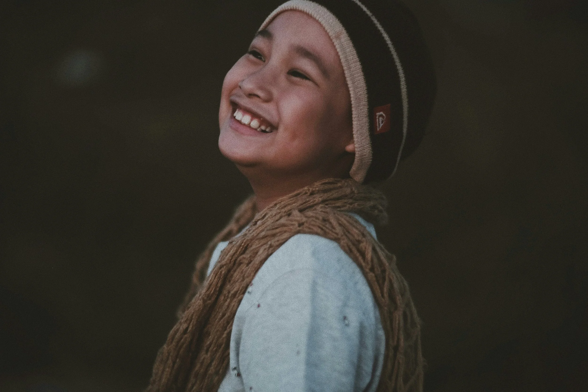 A young boy smiling widely, wearing a knit hat and a jacket with a fleece lining, with dark background.