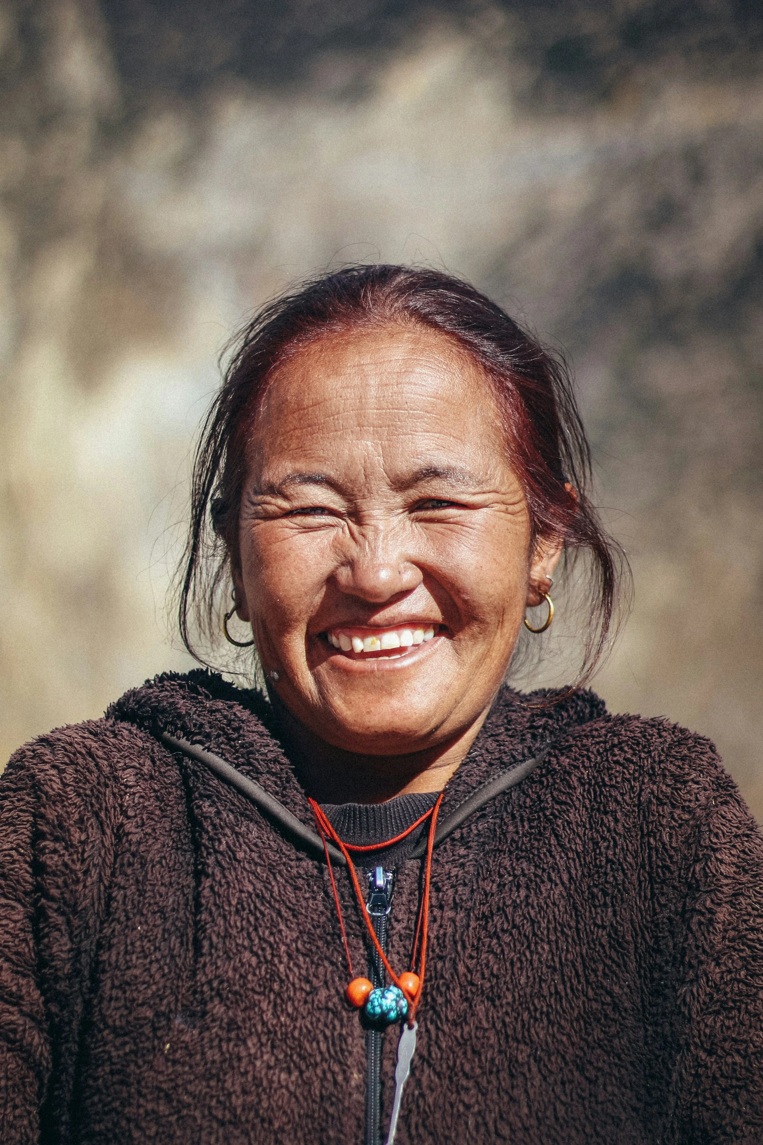 A smiling woman with dark hair tied back, wearing hoop earrings, a maroon fleece jacket, and a beaded necklace, outdoors with a blurry background.