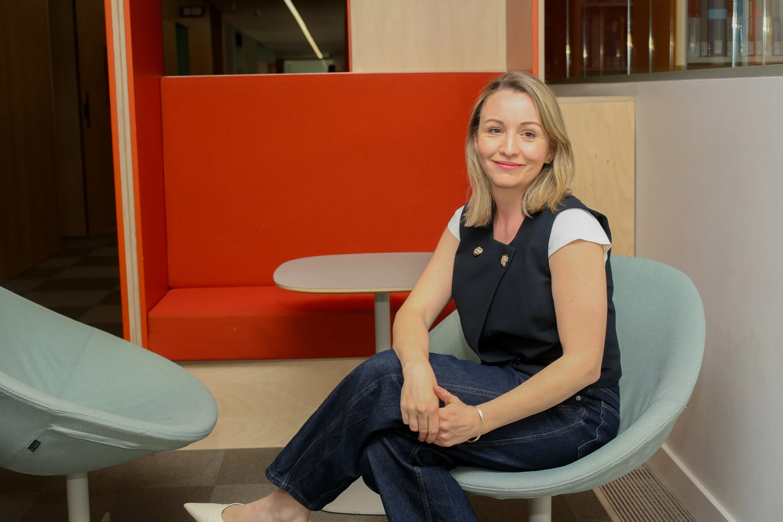 A smiling woman with blonde hair, wearing a black vest over a white shirt, sitting on a modern light green chair in a colorful, modern office space.