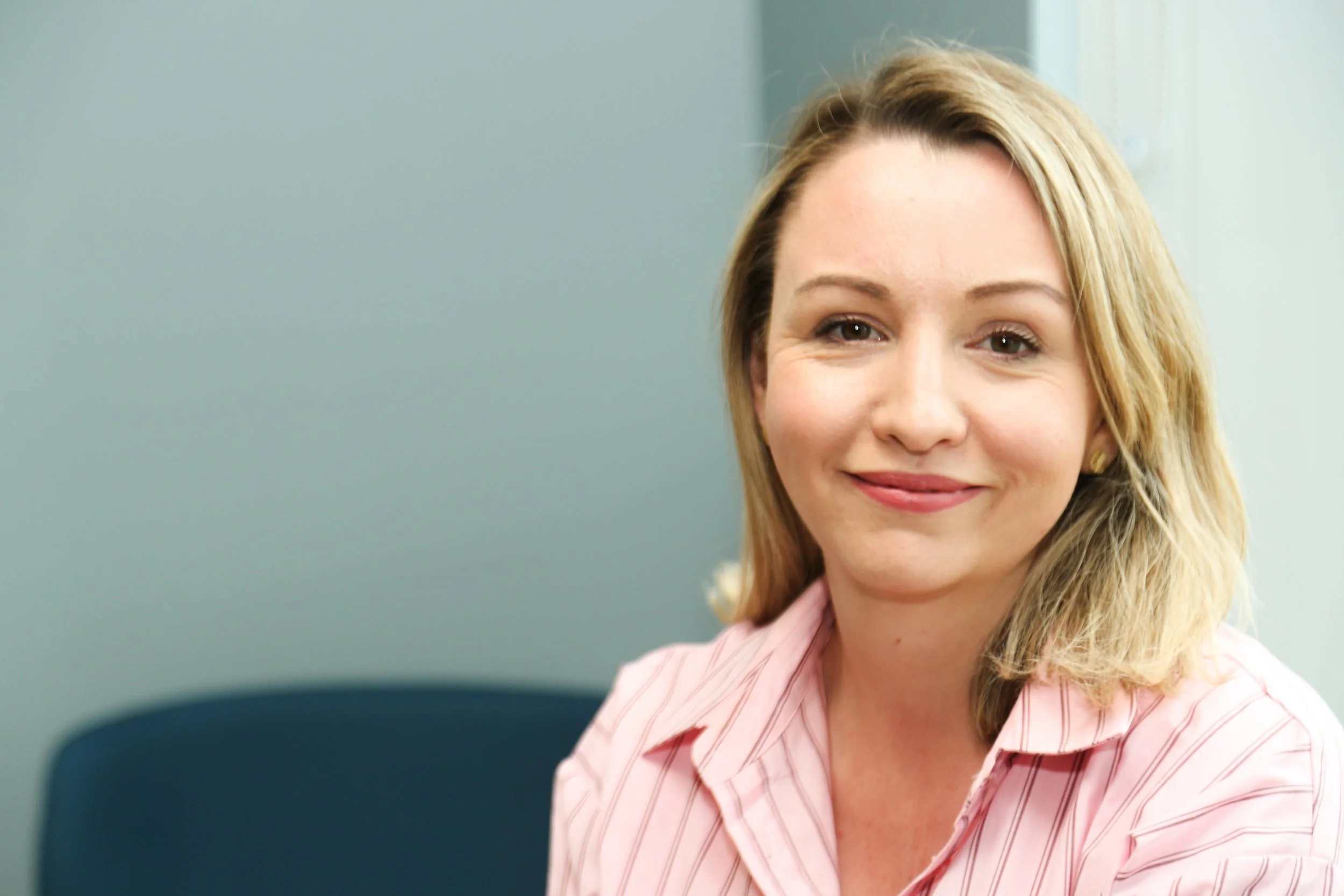 A woman with blonde hair sitting in a chair, smiling, wearing a pink striped shirt.