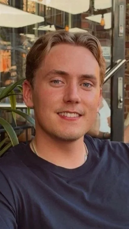 Ung mann med lyst hår og blå øyne smiler i en kafé utendørssetting, med planter og parasoller i bakgrunnen.