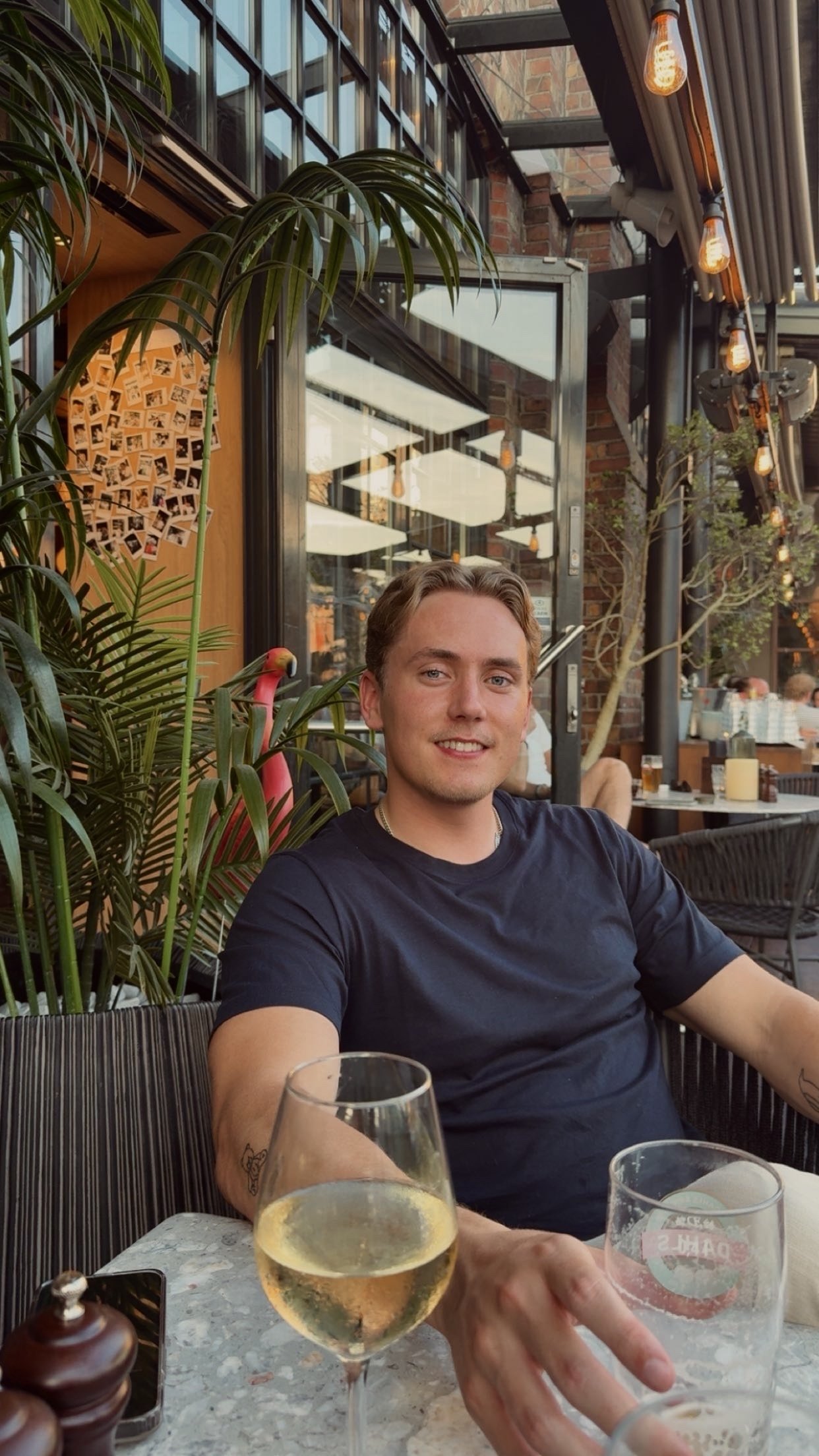 En mann smiler mens han sitter på en utendørs restaurant, omgitt av grønnsaker og lamper, med et glass hvitvin foran seg.