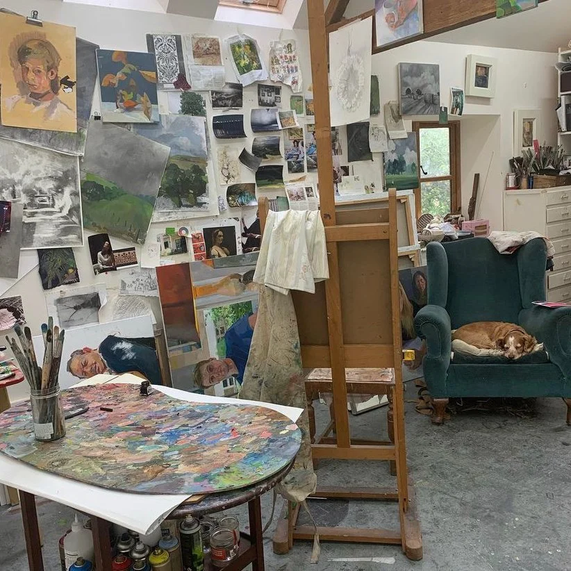 An artist's studio with numerous paintings and sketches on the wall and windows, a colorful palette on the table, a wooden easel, a teal armchair with a sleeping brown and white dog, and art supplies and tools scattered throughout the room.