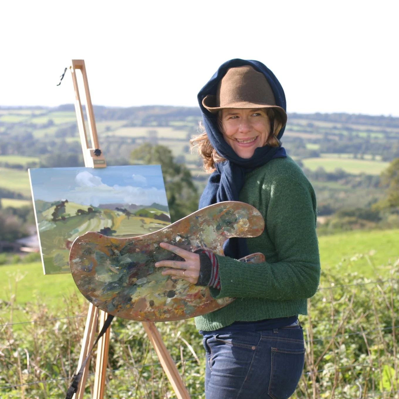 A woman outdoors in a green field, wearing a brown hat, a green sweater, and a scarf, smiling while holding a painter's palette. Behind her, there is an easel with a landscape painting of rolling hills, trees, and clouds.