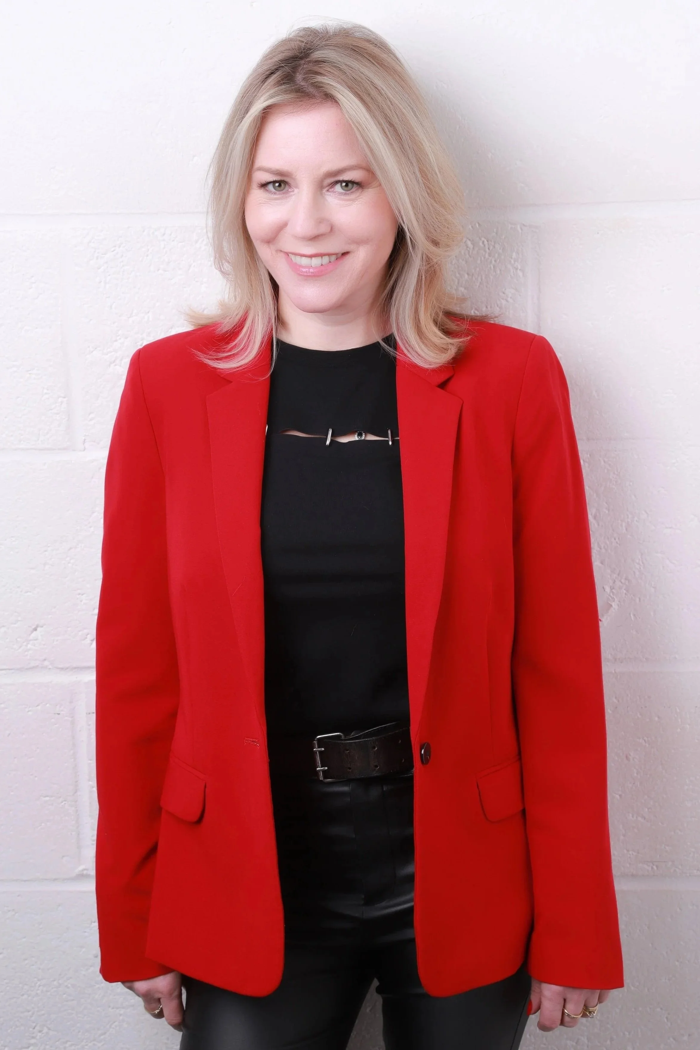 A woman with blonde hair smiling and wearing a red blazer over a black top, standing against a white brick wall.